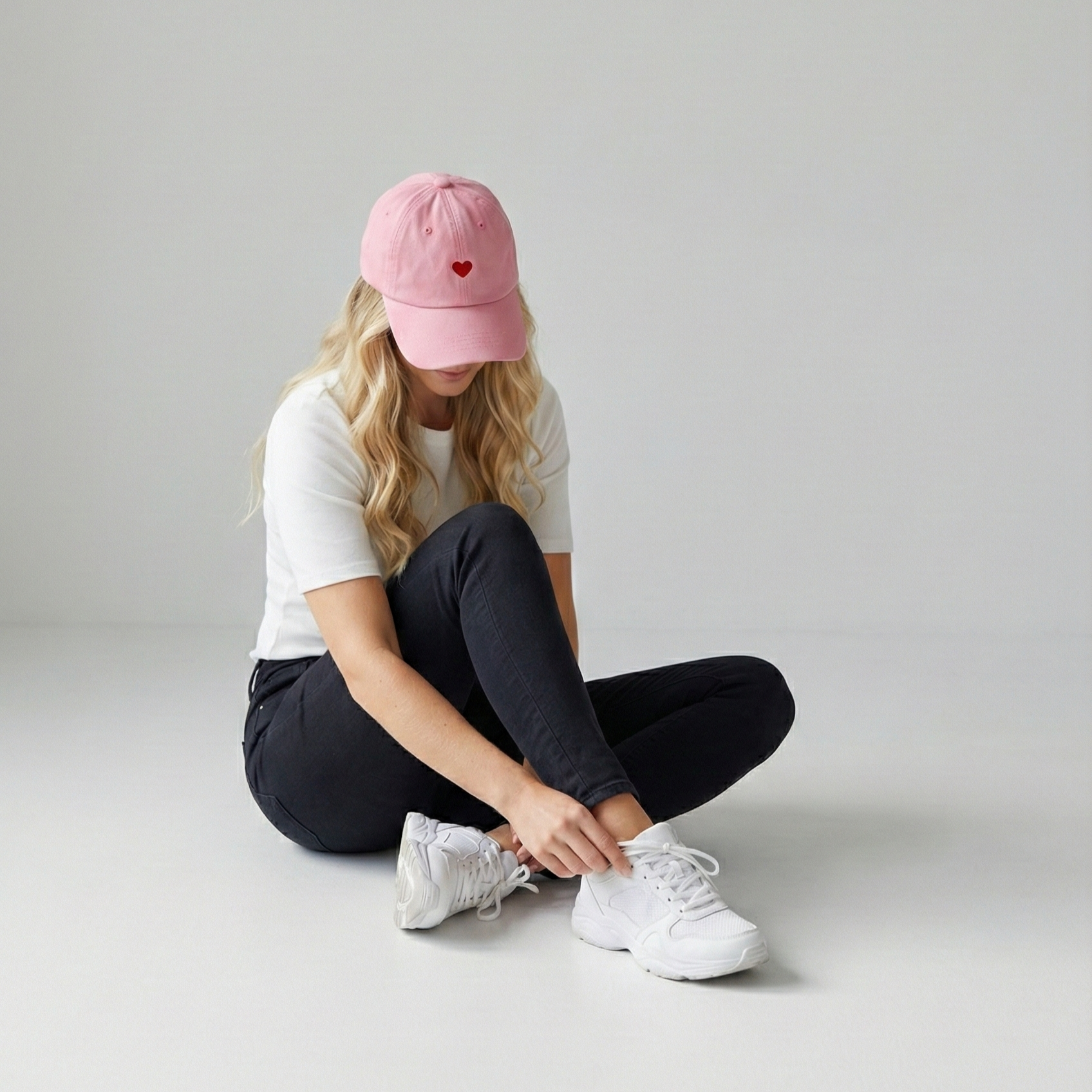 Frau sitzt auf dem Boden, trägt eine weiße T-Shirt, schwarze Hosen, weiße Turnschuhe und eine pinke Kappe mit rotem Herz auf der Vorderseite, während sie ihre Schuhe schnürt.