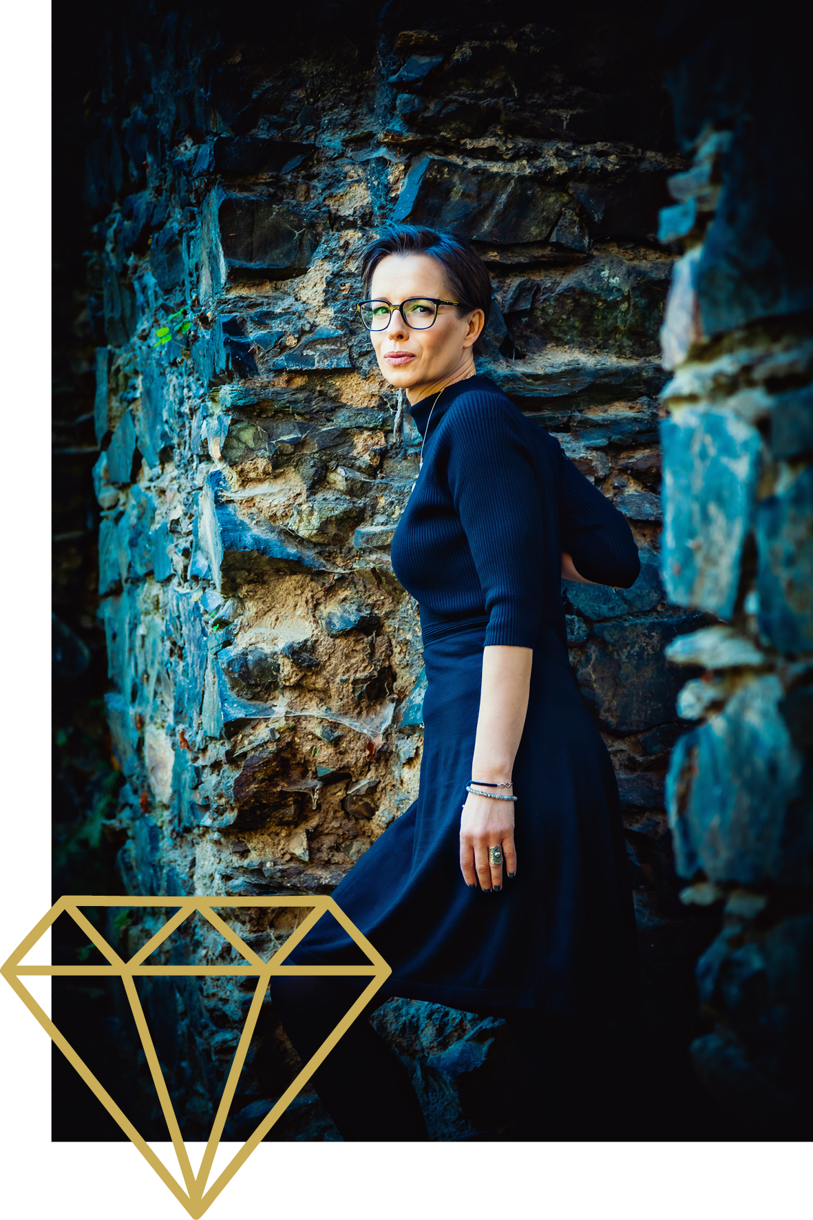 Eine Frau mit kurz schwarzen Haaren, Brille und dunklem Kleid steht vor einer Steinmauer, schaut in die Kamera. Im unteren linken Bildbereich ein goldener Diamantsymbol.