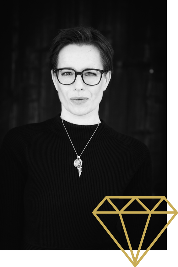 Schwarz-weiß-Fotografie einer Frau mit kurzen Haaren, Brille und Ketten, die direkt in die Kamera blickt. Im unteren rechten Eck ein goldener Diamanten-Icon.