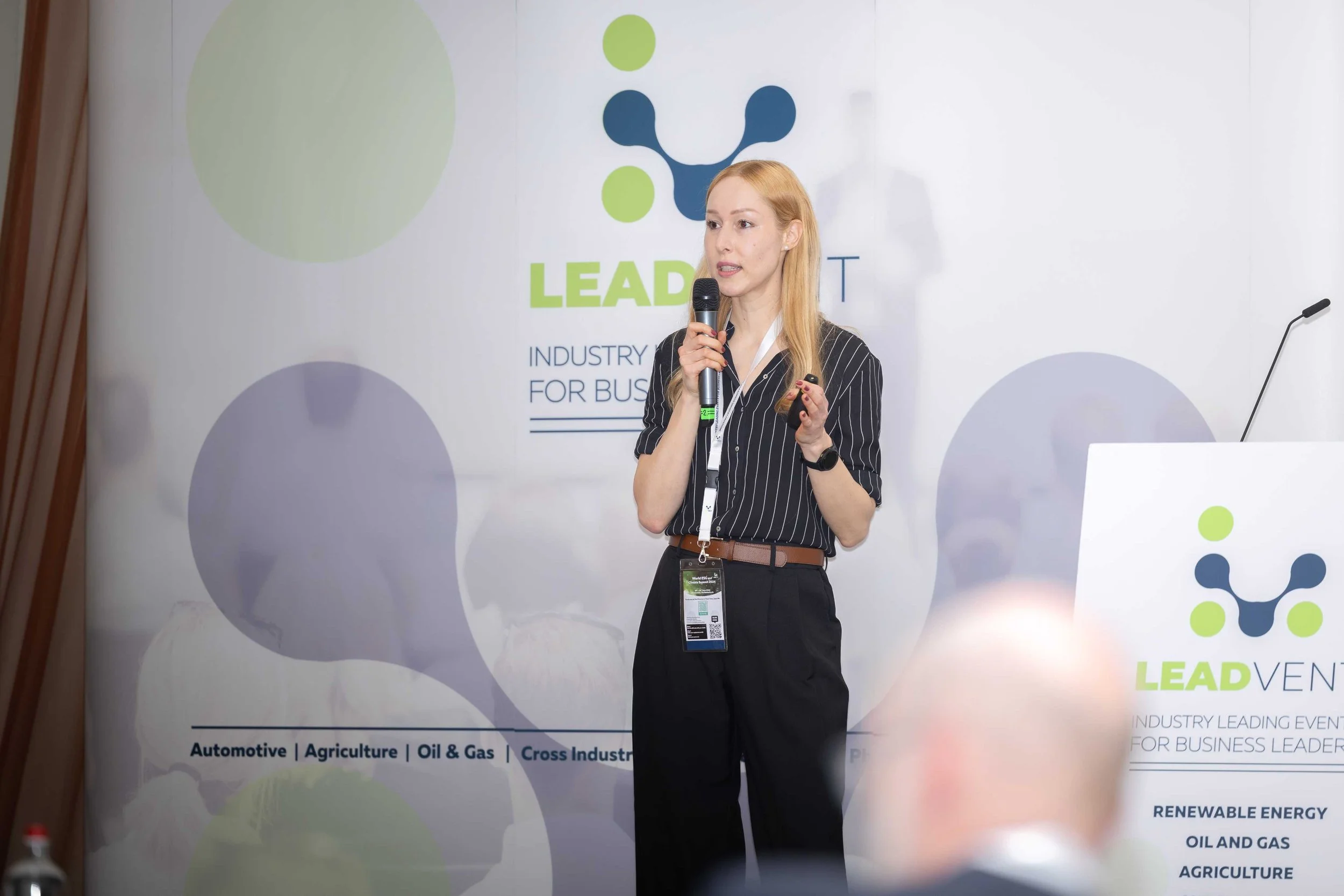 A woman with long blonde hair giving a presentation at a conference, holding a microphone. She is standing in front of a large banner with the words 'LEADVENT' and a colorful logo. The banner includes topics like automotive, agriculture, oil & gas, a