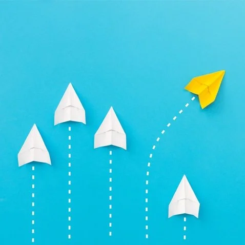 Paper airplanes against blue background, with four white planes moving straight and one yellow plane veering off in a different direction.