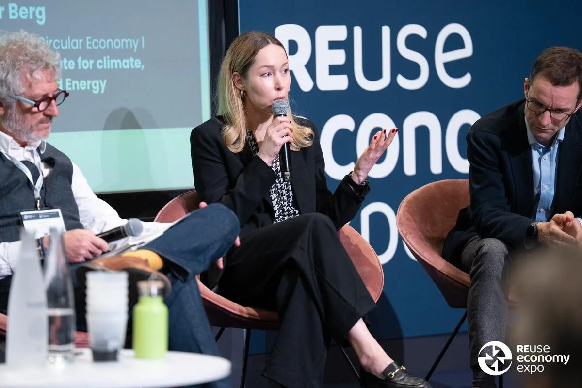 Three individuals seated on stage at a panel discussion during the Reuse Economy Expo. The woman in the center is speaking into a microphone, wearing a black blazer and trousers. The man on the left has gray hair, glasses, and is holding a device, while the man on the right is dressed in a dark blazer and glasses, looking down. The background features a blue screen with the words 'REUSE' and 'CONC' visible, and the expo logo in the lower right corner.