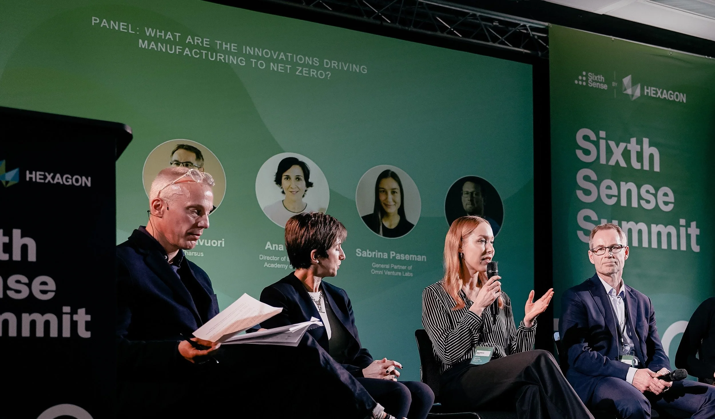 A panel discussion at the Sixth Sense Summit with four people on stage, one woman speaking into a microphone, and a green backdrop displaying event branding and speaker names.