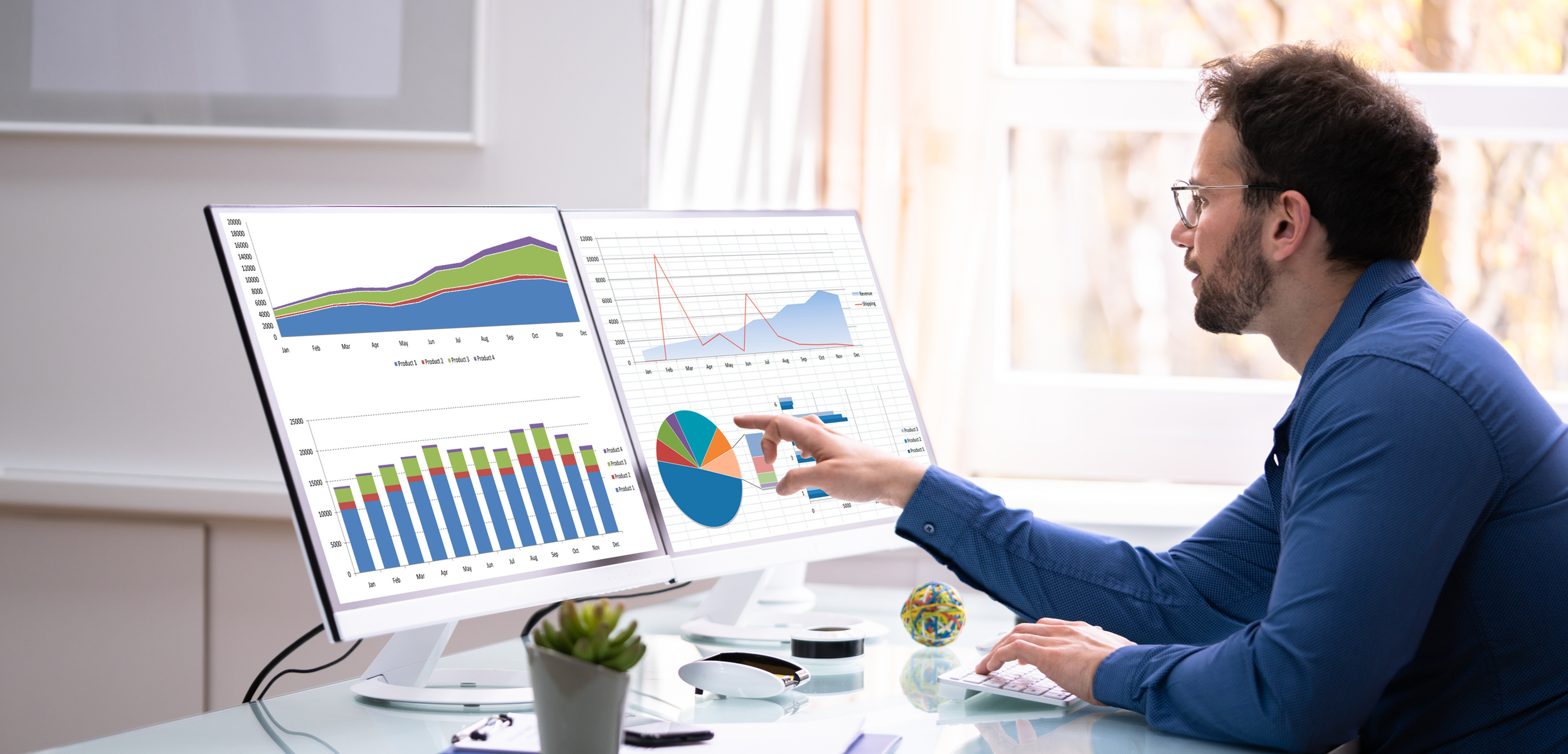Man in a blue shirt analyzing graphs and charts on dual monitors, using a keyboard and mouse in an office setting.