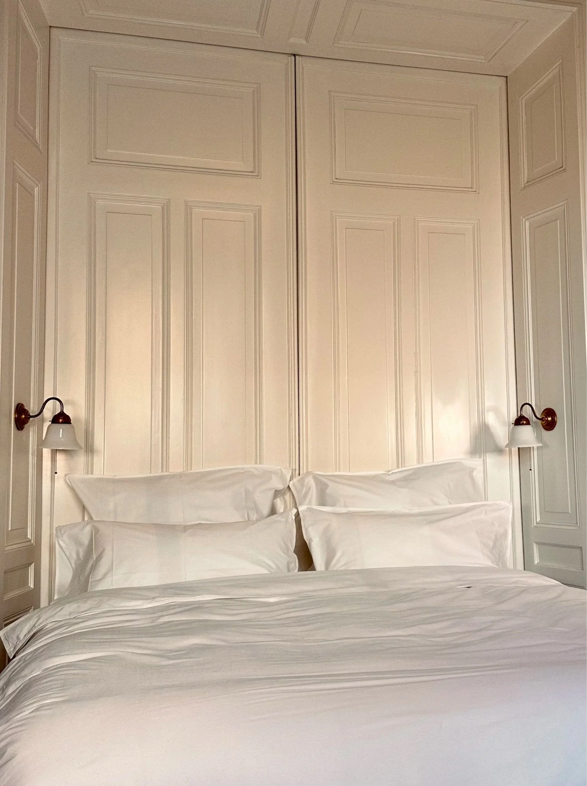 A neatly made bed with white pillows and bedding, flanked by wall-mounted lamps on either side, against a background of cream-colored paneled walls.