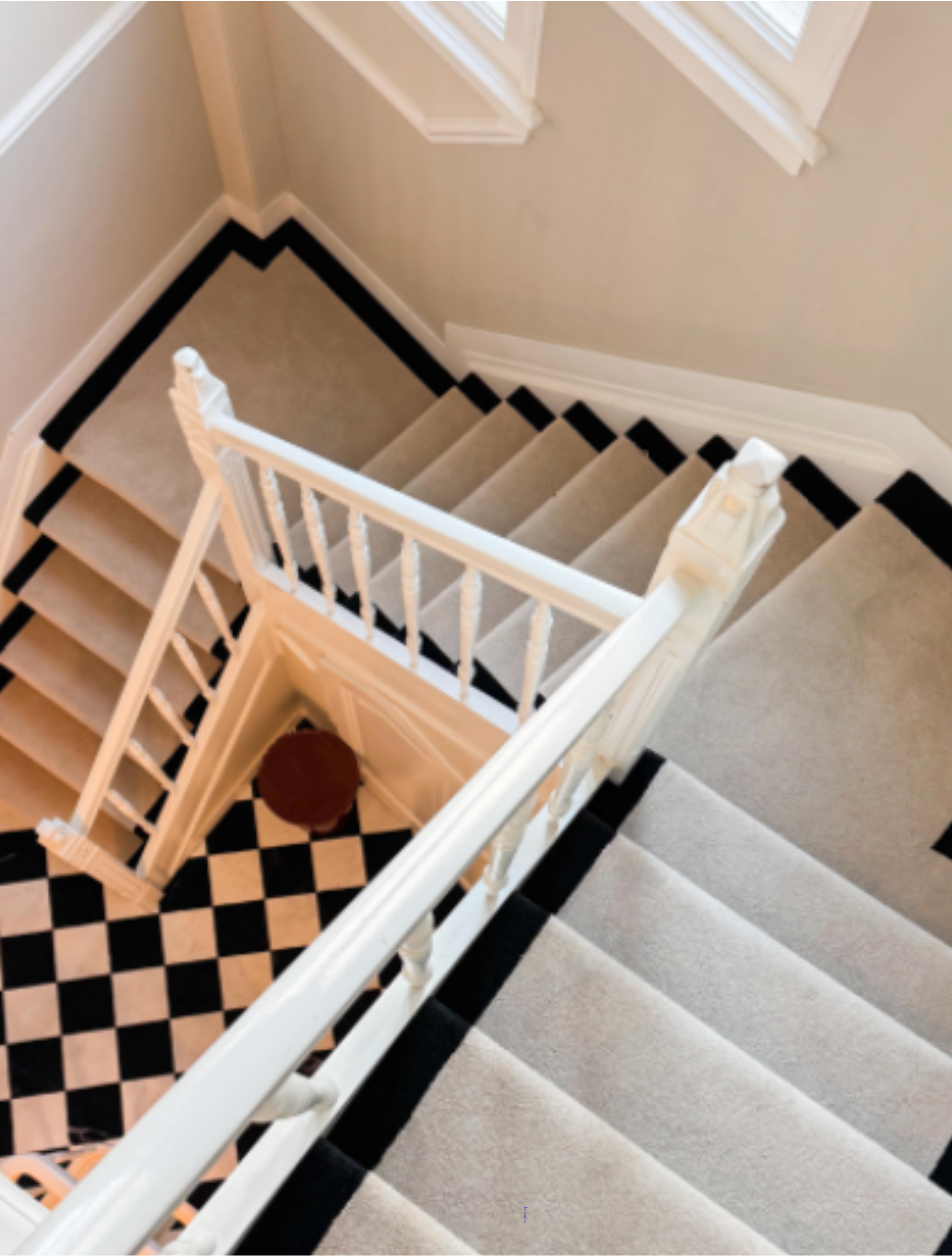 View looking down at a staircase from above, showing the black and white checkered floor entryway and beige carpeted stairs with a white railing.