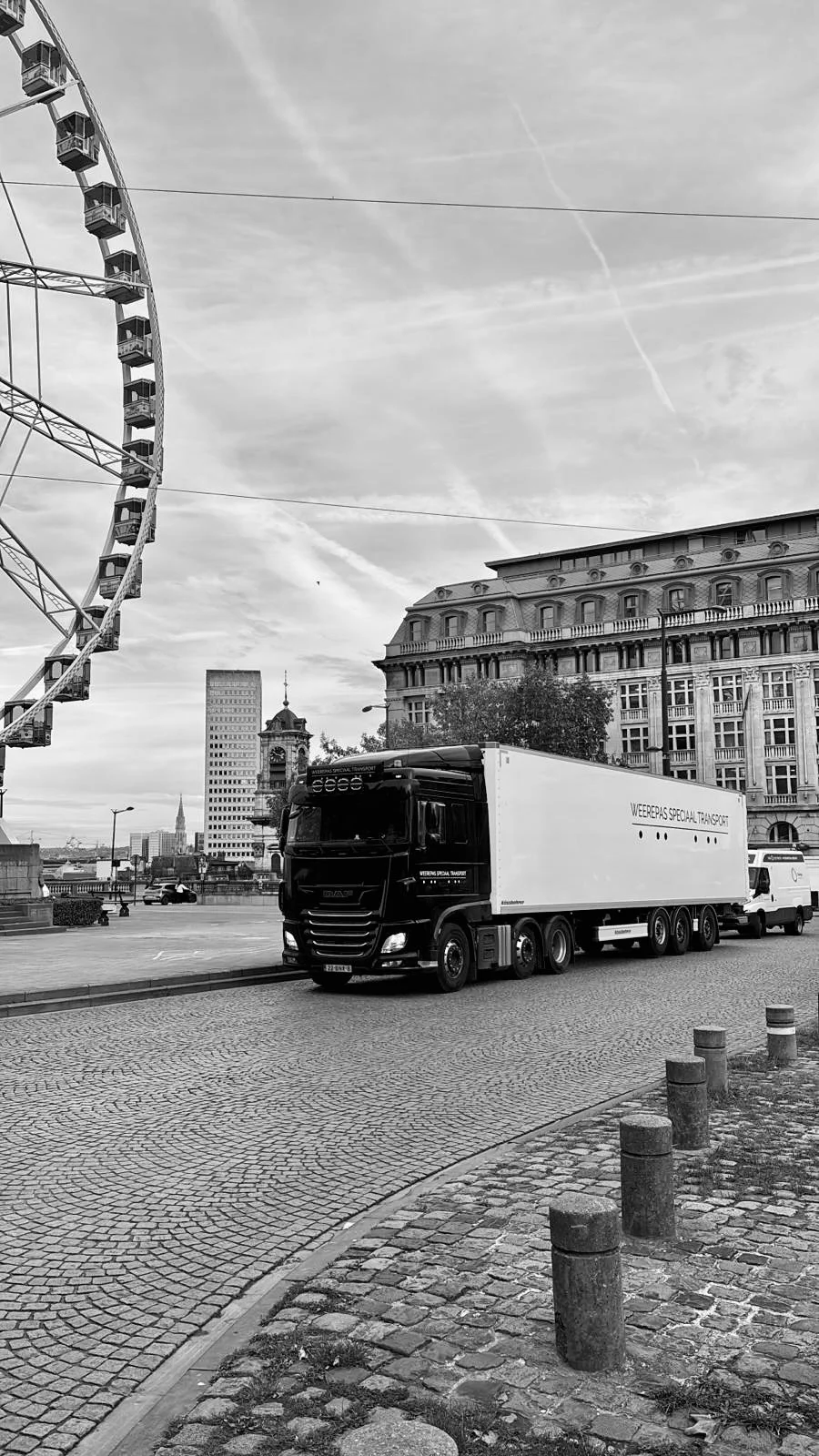 A city street scene with a large ferris wheel on the left, a truck and a van parked on the right, historic buildings in the background, and a cloudy sky overhead.