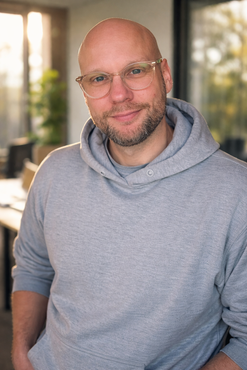 Ein lächelnder Mann mit Brille, Bart und Glatze trägt einen grauen Hoodie, im Hintergrund ein modernes, hell erleuchtetes Büro mit Fenstern und Pflanzen.