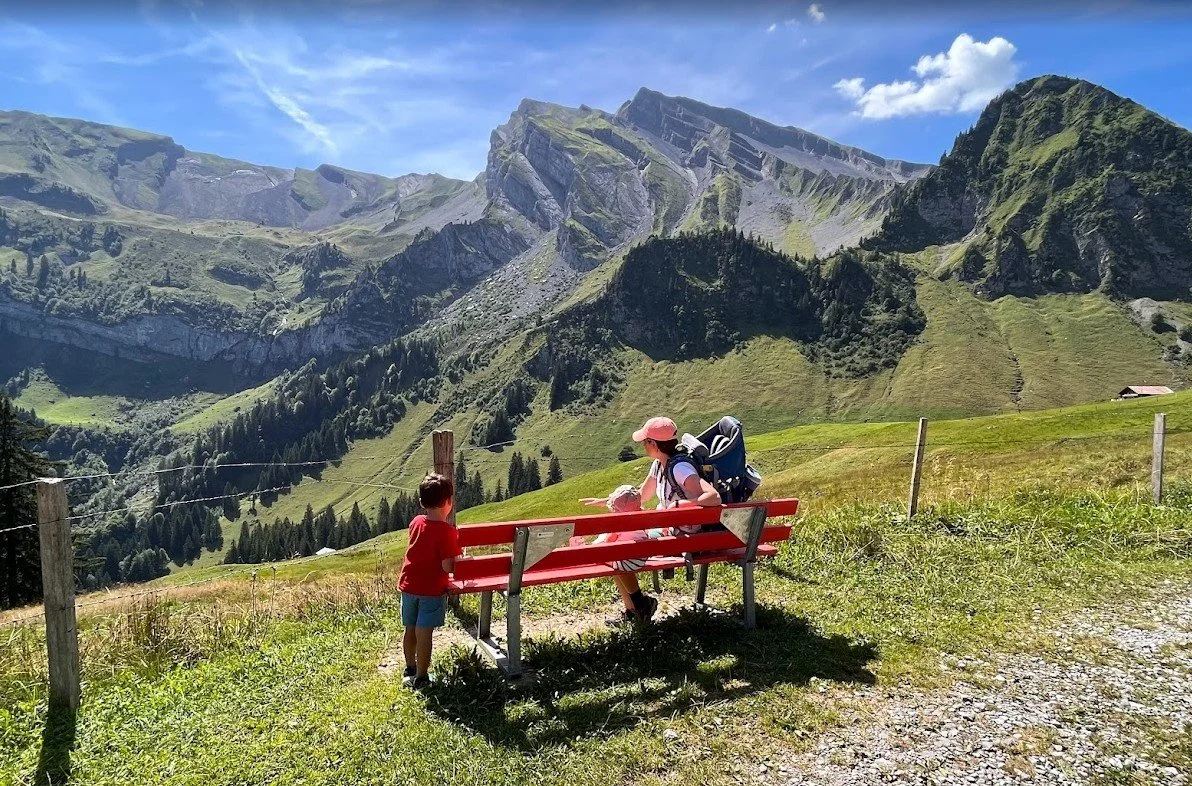 A woman and a young boy sitting on a red bench outdoors in a mountain landscape. The woman is wearing a pink hat and sunglasses, with a backpack, and is pointing at the scenery. The boy is standing next to the bench, wearing a red shirt and blue shorts, and looking at the woman. The background features green grassy hills and rugged mountains under a partly cloudy sky.