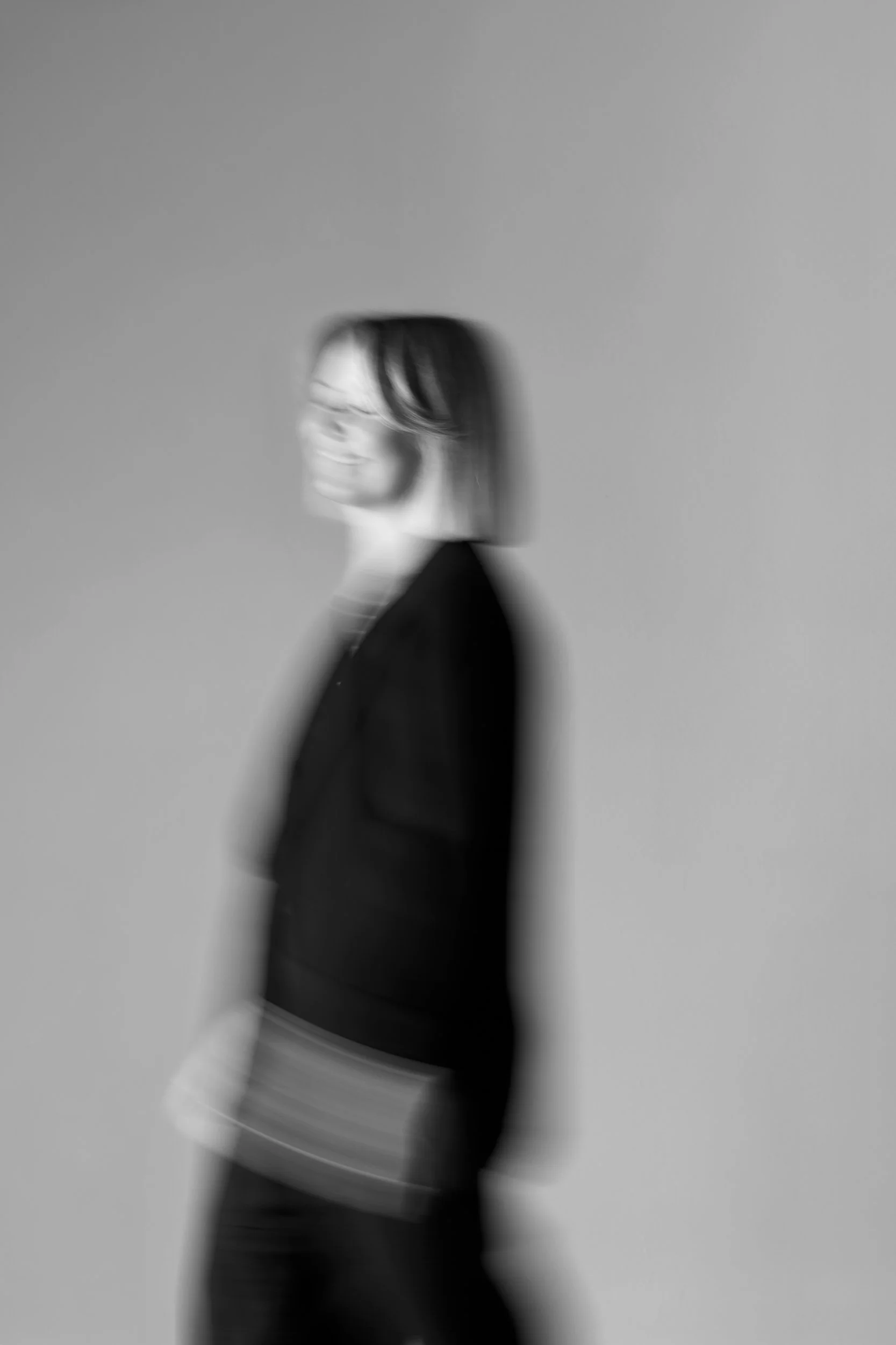 Sort-hvid portrætfoto af en smilende kvinde med brunt hår, iført sort jakke og tøj, fotograferet mod en neutral grå baggrund.