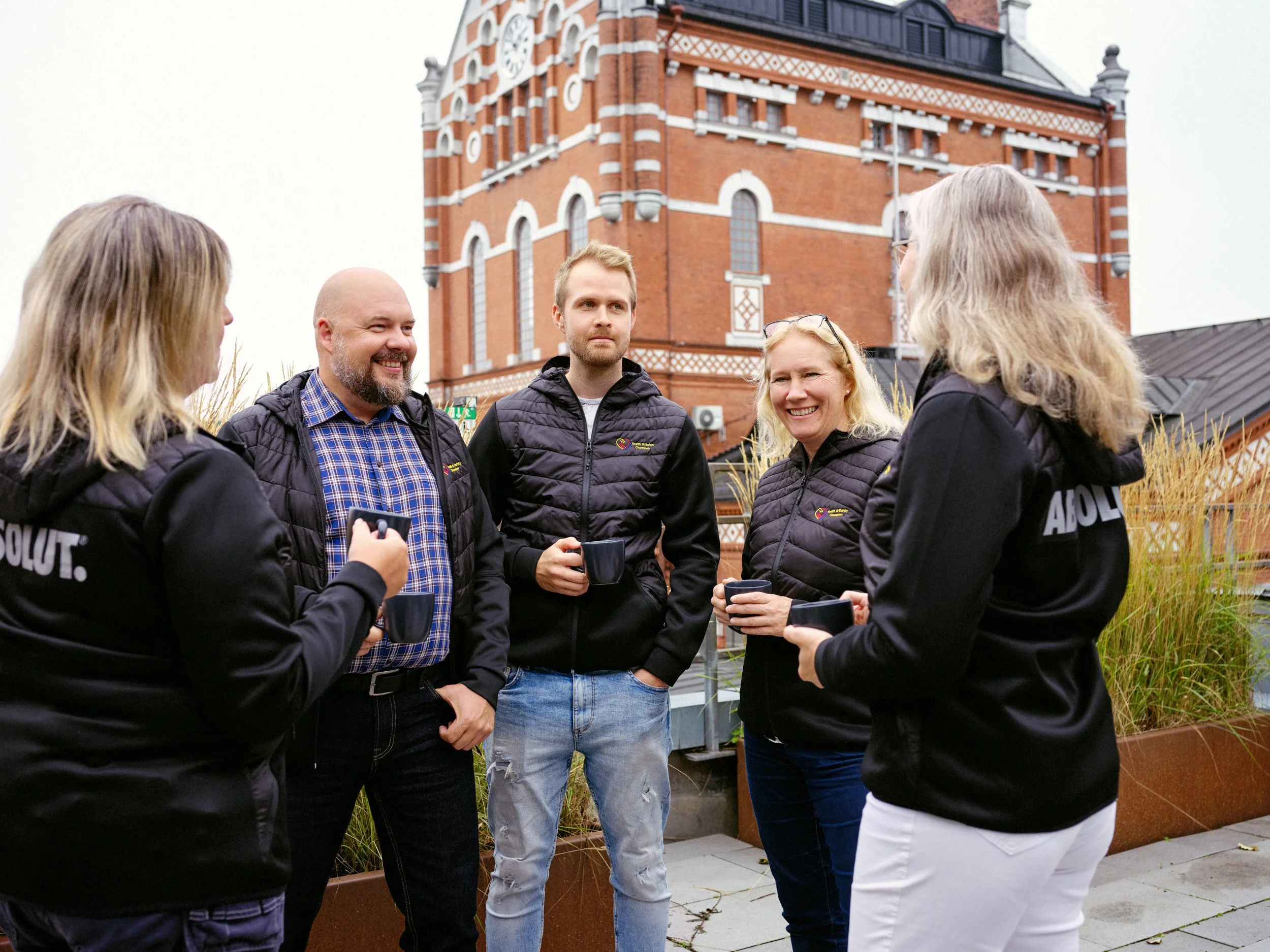 En grupp människor står utomhus framför en gammal tegelbyggnad. De verkar konversera och hålla kaffekoppar. Personerna är klädda i svarta jackor och det finns grönska i bakgrunden.