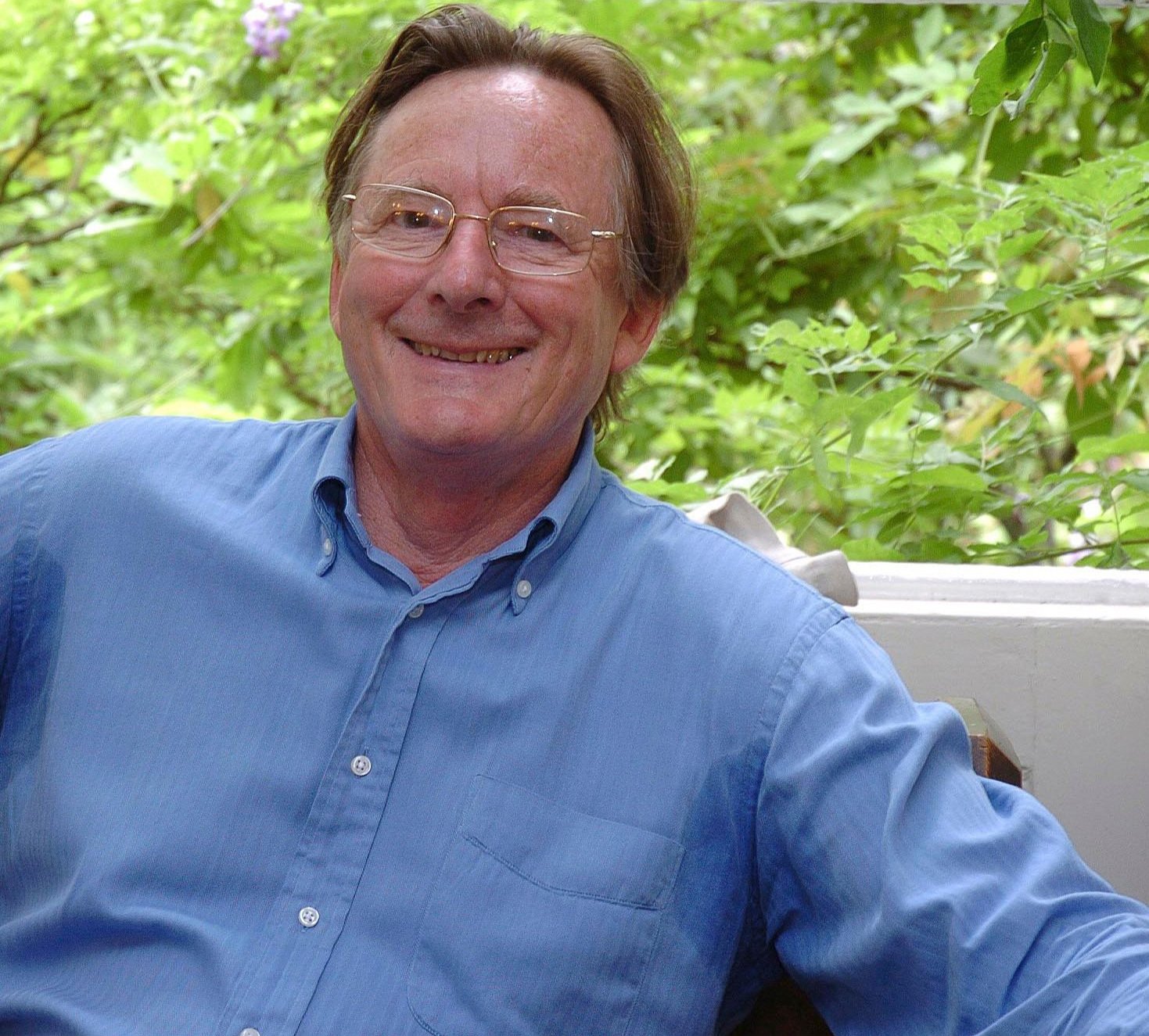A smiling  John Carewe aged 70 wearing glasses and a light blue button-up shirt, sitting outdoors with green foliage in the background.