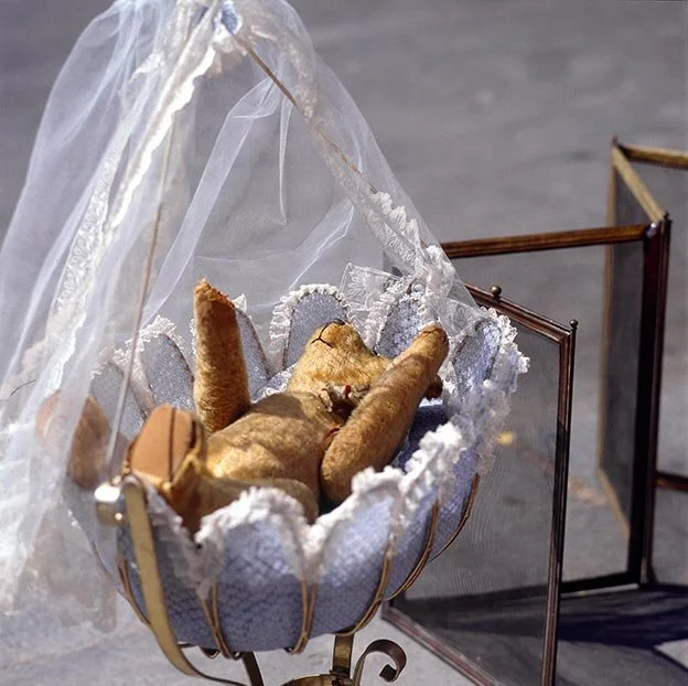 A dog lying on its back in a small, ornate cradle with a sheer canopy, decorated with lace and ribbons.