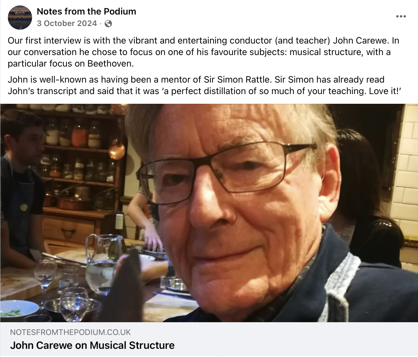 Close-up of John Carewe wearing glasses at a dining table with glasses and bowls, in a cozy restaurant setting, with other people and wooden shelves in the background.
