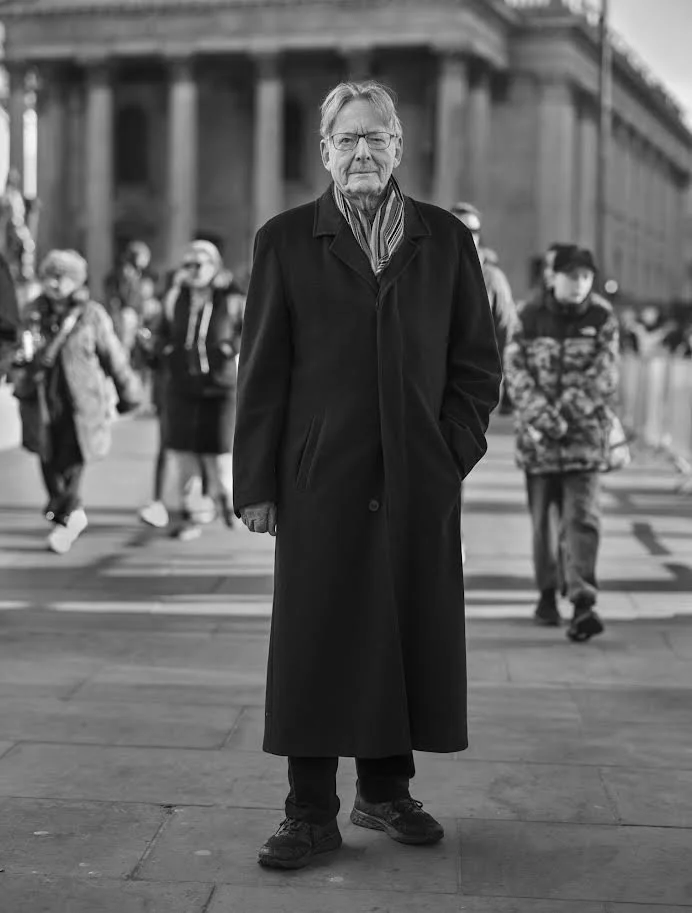John Carewe aged 90 wearing a long coat and scarf standing on a city street with people walking in the background, black and white photo.