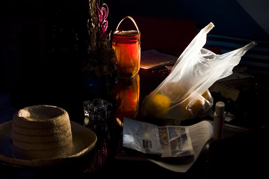A cluttered table with a straw hat, a glass jar with a candle inside, a plastic bag with lemons, a magazine, a white bottle, and a lit candle in the background.