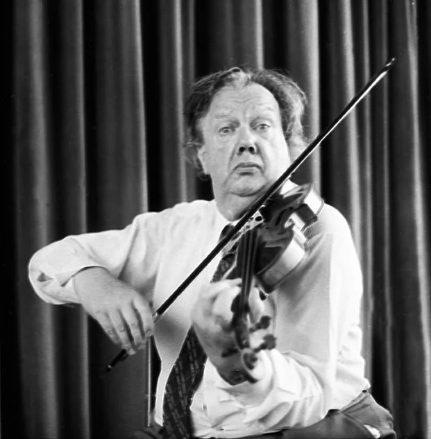 Black and white photo of a man playing a violin with a serious expression, standing in front of a curtain.