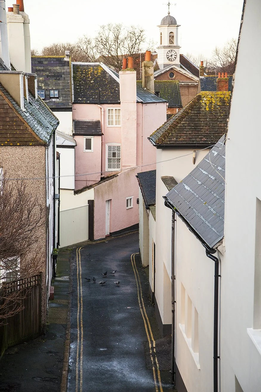 A narrow alleyway between colorful buildings with a small group of pigeons on the wet pavement. In the background, there is a church clock tower with a weather vane on top.
