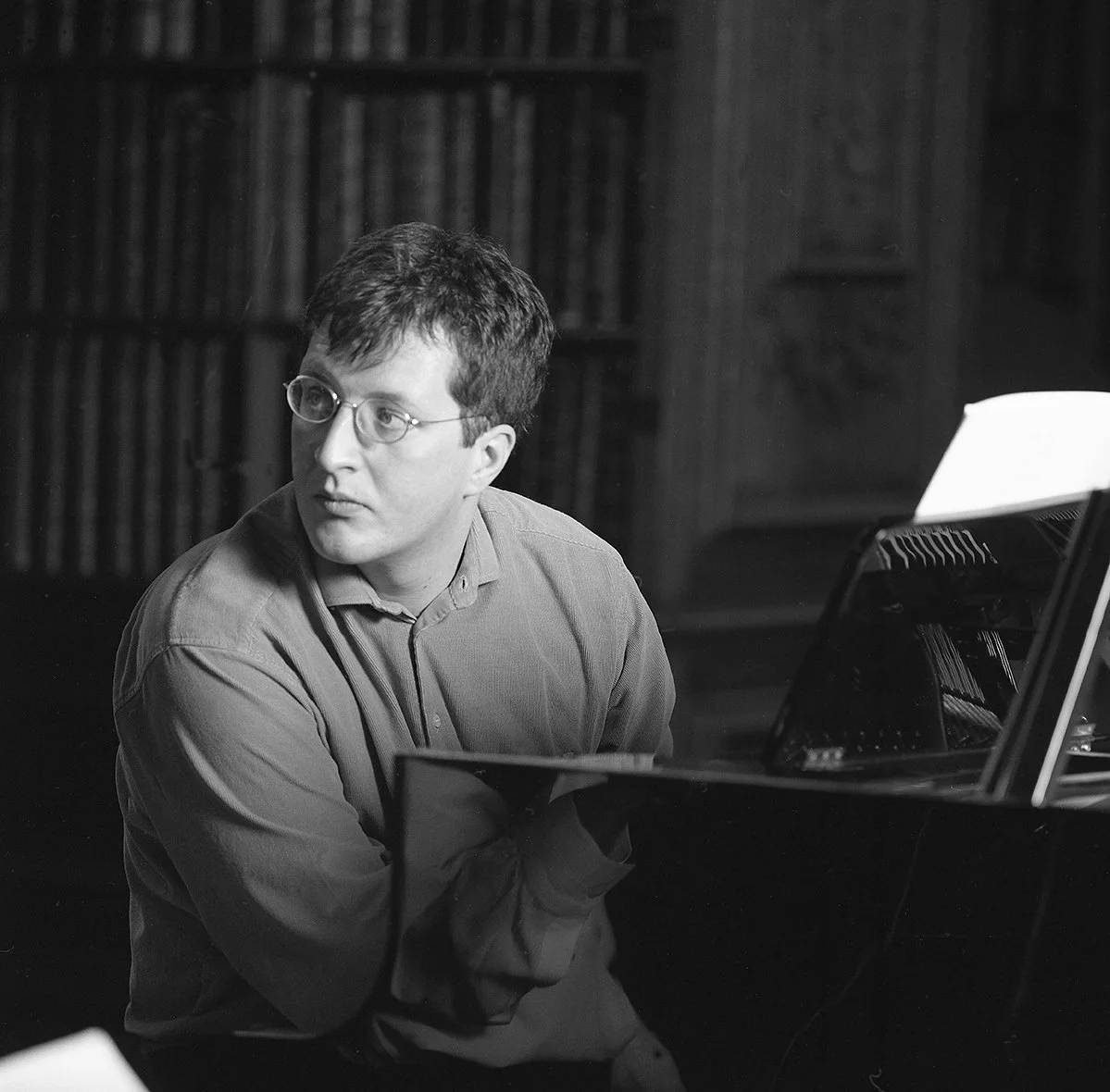 Image of a young Thomas Ades with short, dark hair and glasses sitting at a piano, looking off to the side with a focused expression. He is wearing a collared shirt and is in a room with bookshelves in the background.
