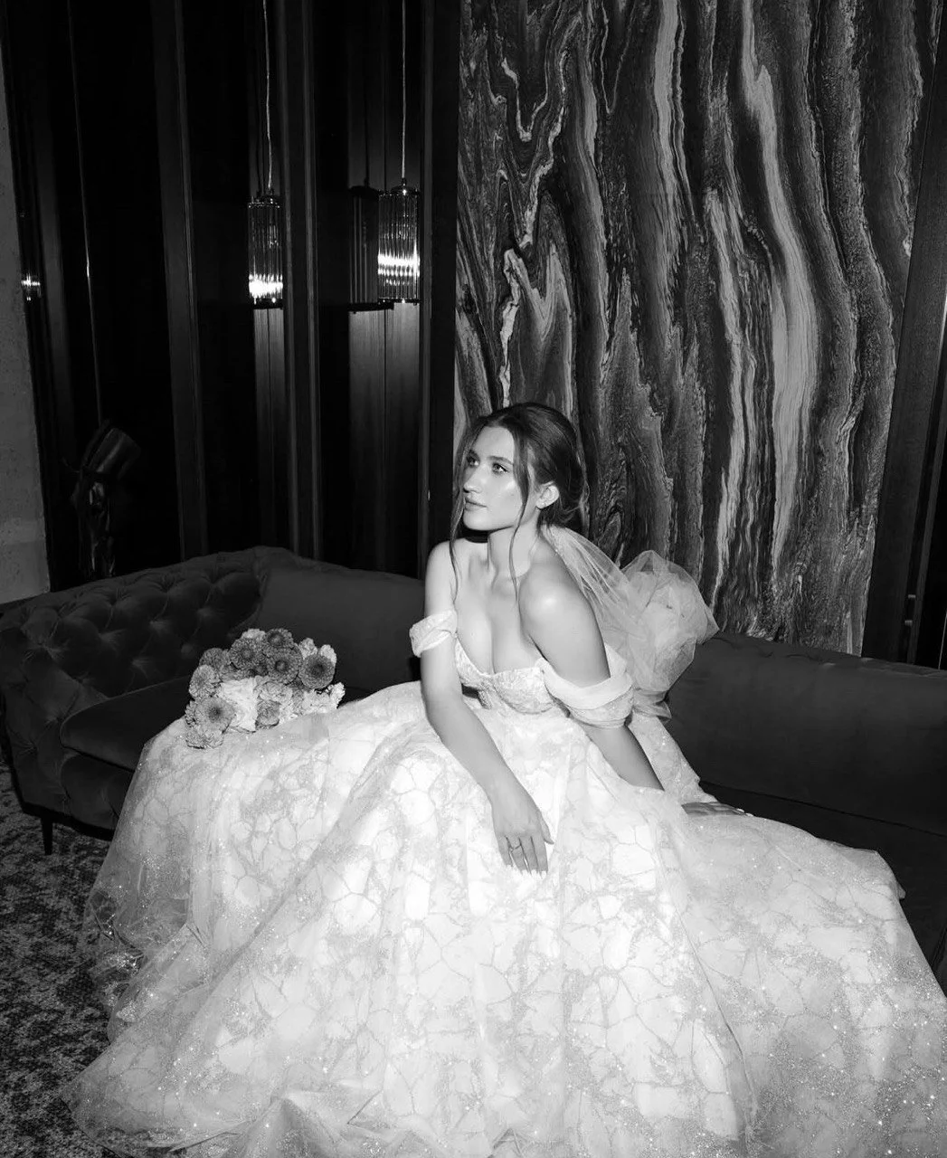 Black and white photo of a woman in a luxurious off-shoulder wedding dress, sitting on a plush couch with a bouquet of flowers. She is in an elegant room with intricate wall patterns.