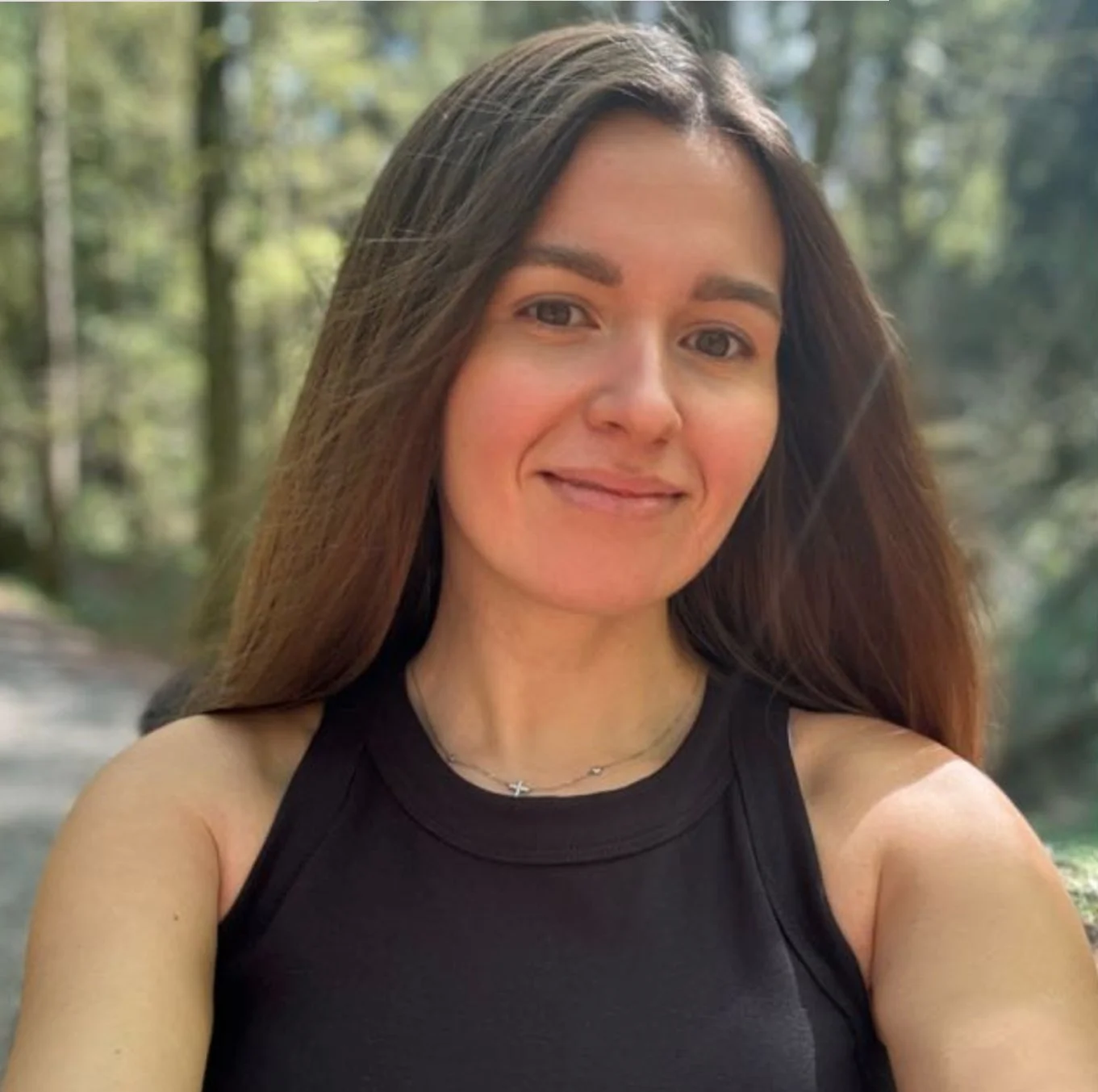 A woman with long hair smiles outdoors, wearing a black sleeveless top. Trees and a pathway are in the background.