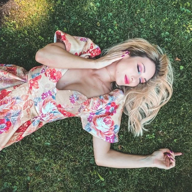 Woman in floral dress lying on grass, eyes closed, hand on face