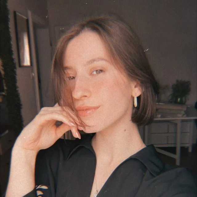 Woman with short brown hair and hoop earrings posing indoors, wearing a black top, with hand resting on her face.