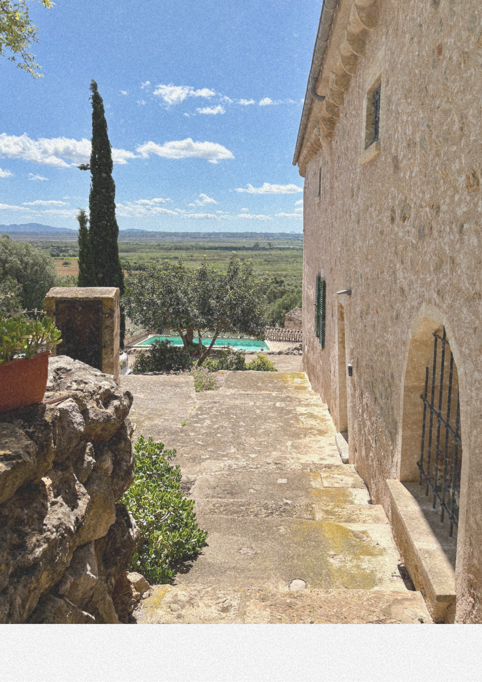 Rustic stone house exterior with stone steps leading to a garden and swimming pool; tall cypress tree and landscape view with blue sky and clouds.