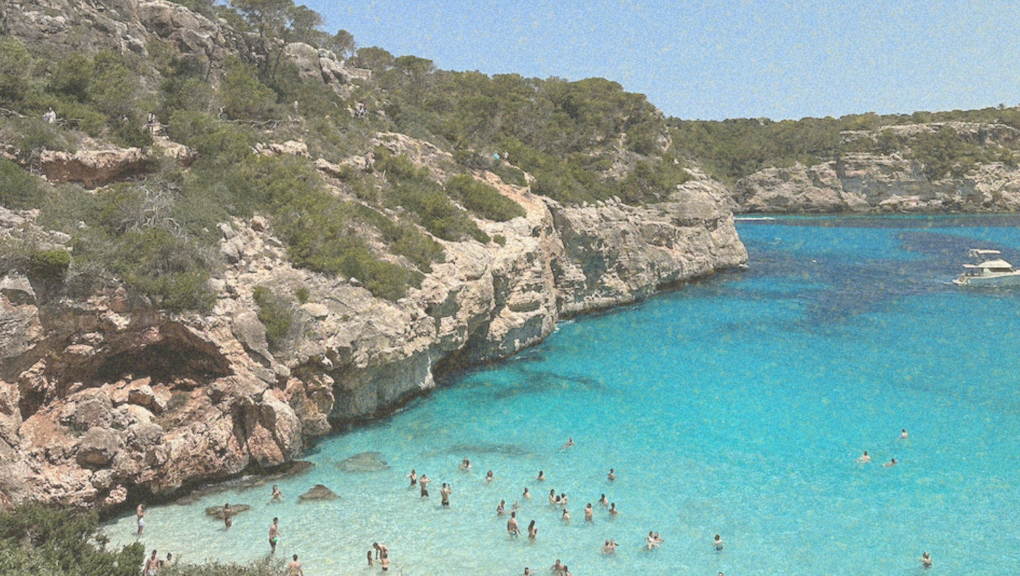 People swimming in clear blue water near rocky cliffs and trees, with a boat in the background.