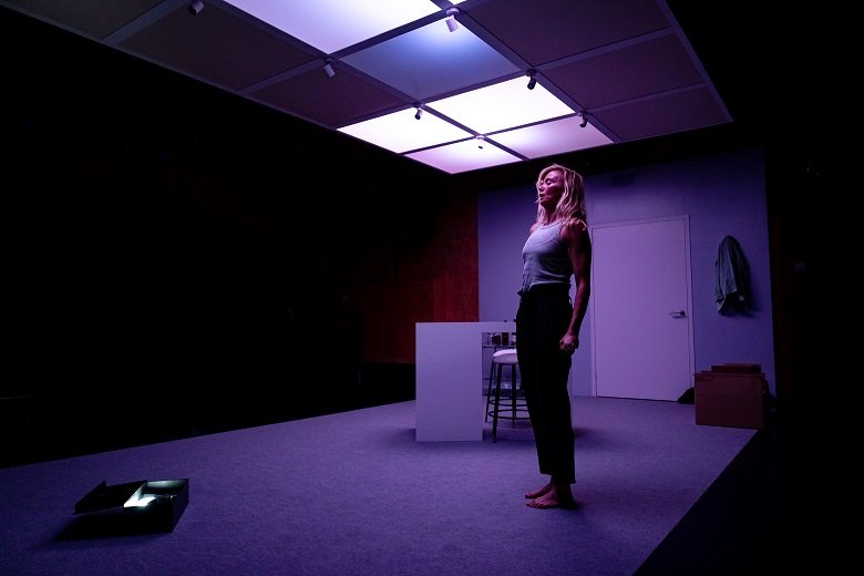 Person standing in a dimly lit room with a modern grid ceiling, wearing casual clothing, and boxes present on the floor.