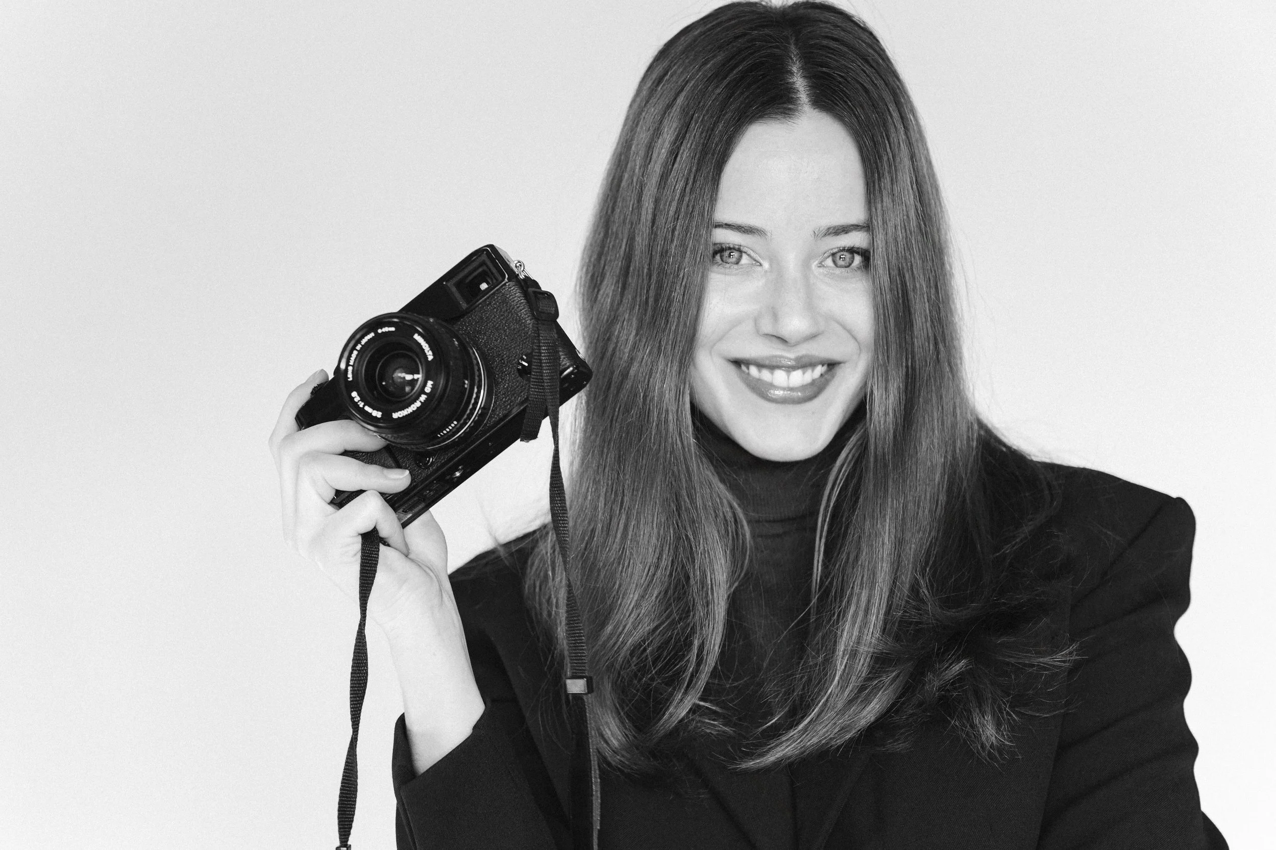 Eine lächelnde Frau mit langem Haar hält eine Kamera in der Hand. Sie trägt einen schwarzen Blazer und vor einem hellen Hintergrund. Portrait einer Fotografin.