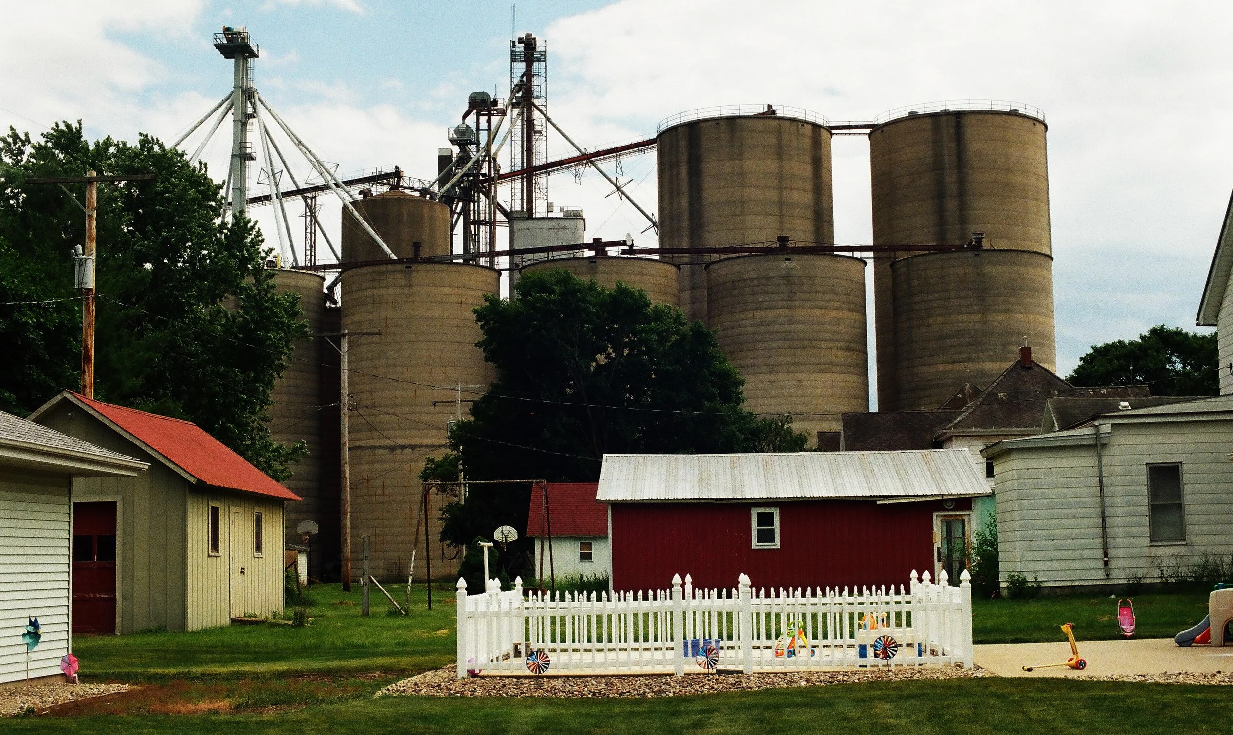 Residential backyard with small houses, lawn, and playground equipment in the foreground, and industrial grain silos and structures in the background under cloudy sky.