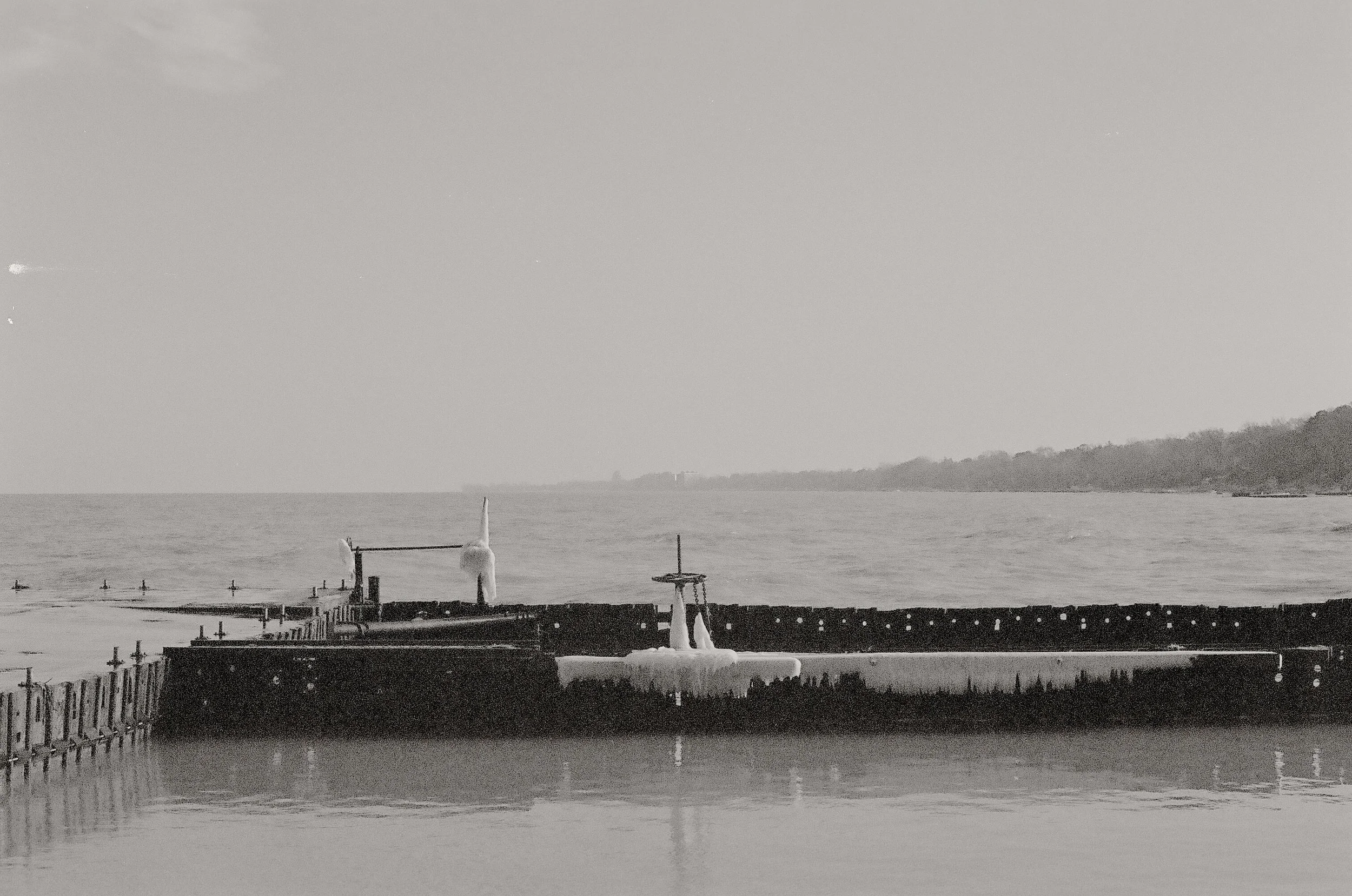 Black and white photo of an ice-covered pier extending into a calm body of water, with a shoreline and trees in the distance under a clear sky.