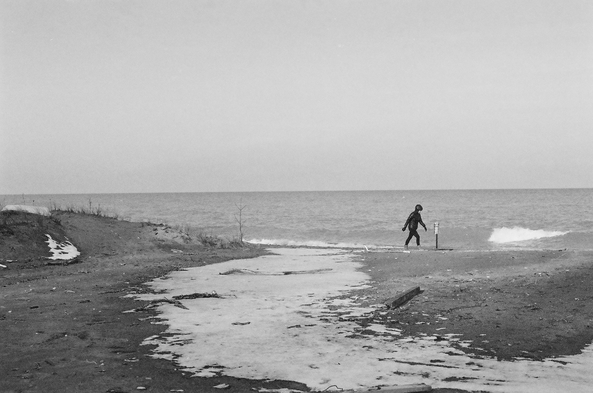 A person wearing a jacket and hat walking on a sandy beach near the ocean with a trail of snow or ice on the ground, in black and white.