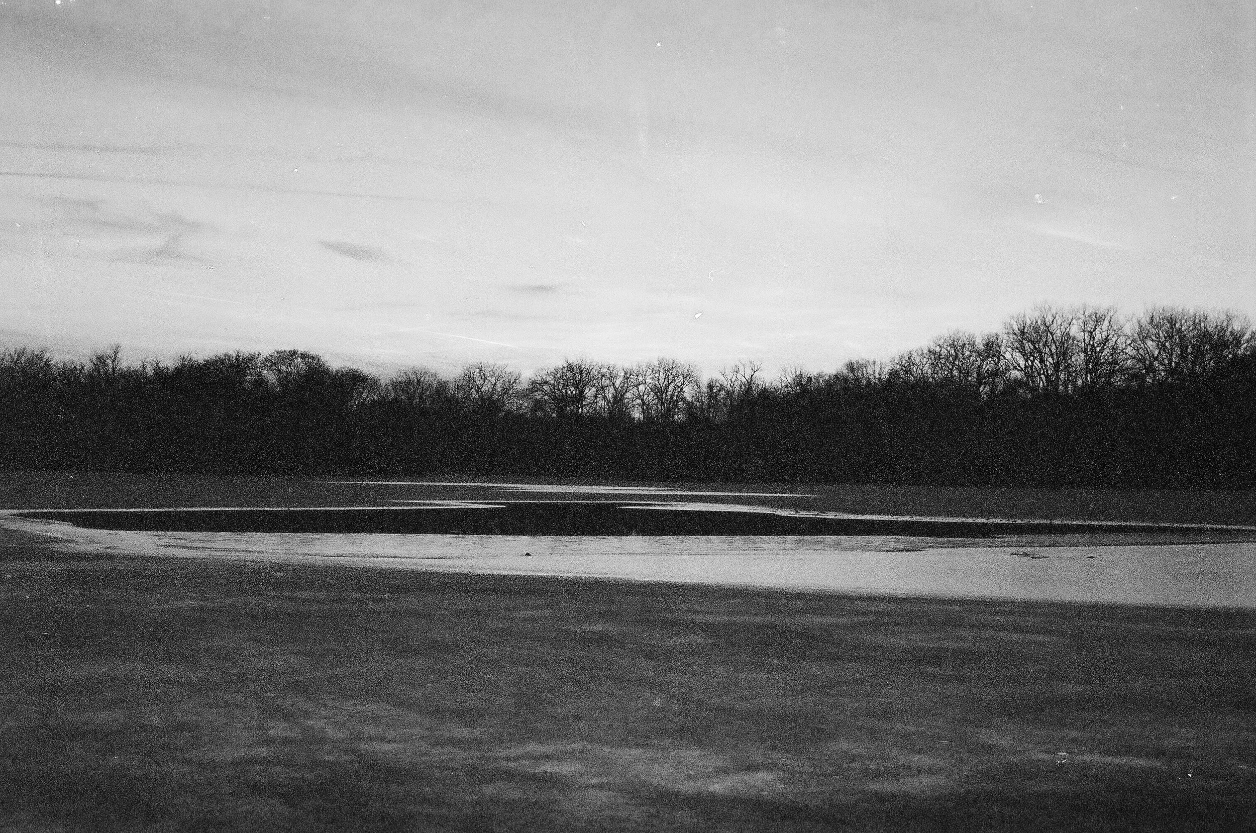 Black and white landscape photo of a frozen lake or river with bare trees in the background and a cloudy sky.