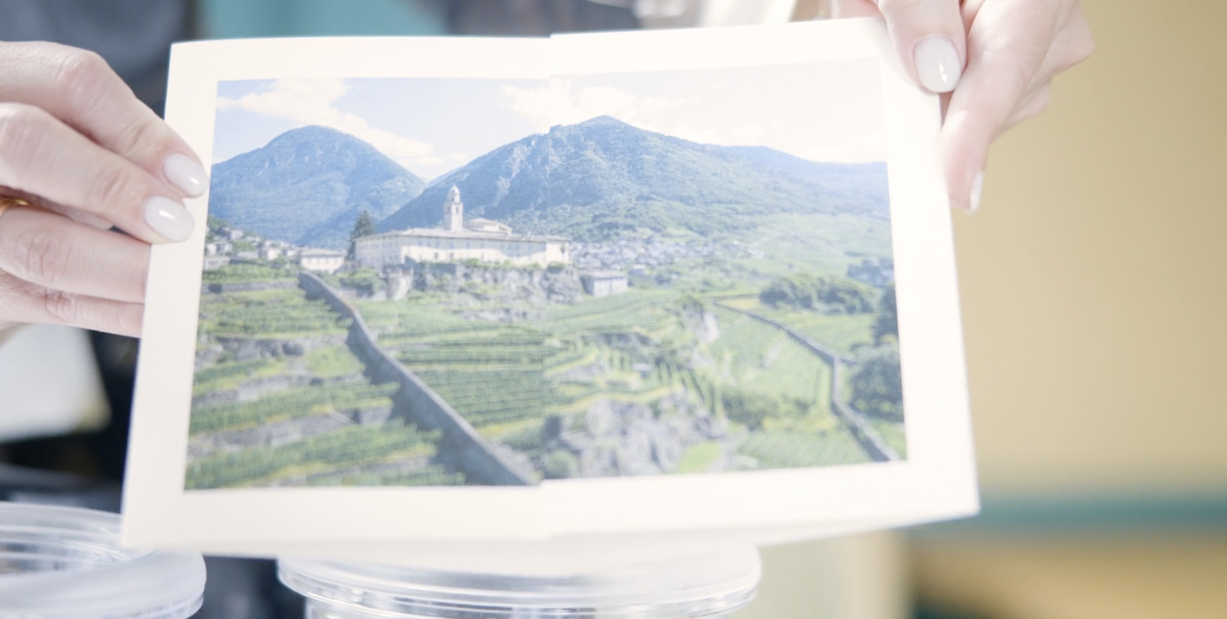 Person holding a printed photo of a landscape featuring terraced vineyards, a building with a clock tower, and mountains in the background.