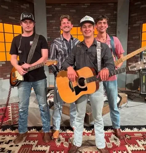 Four young men with musical instruments posing in a studio, two holding guitars, one with a bass guitar, and one in the back smiling.