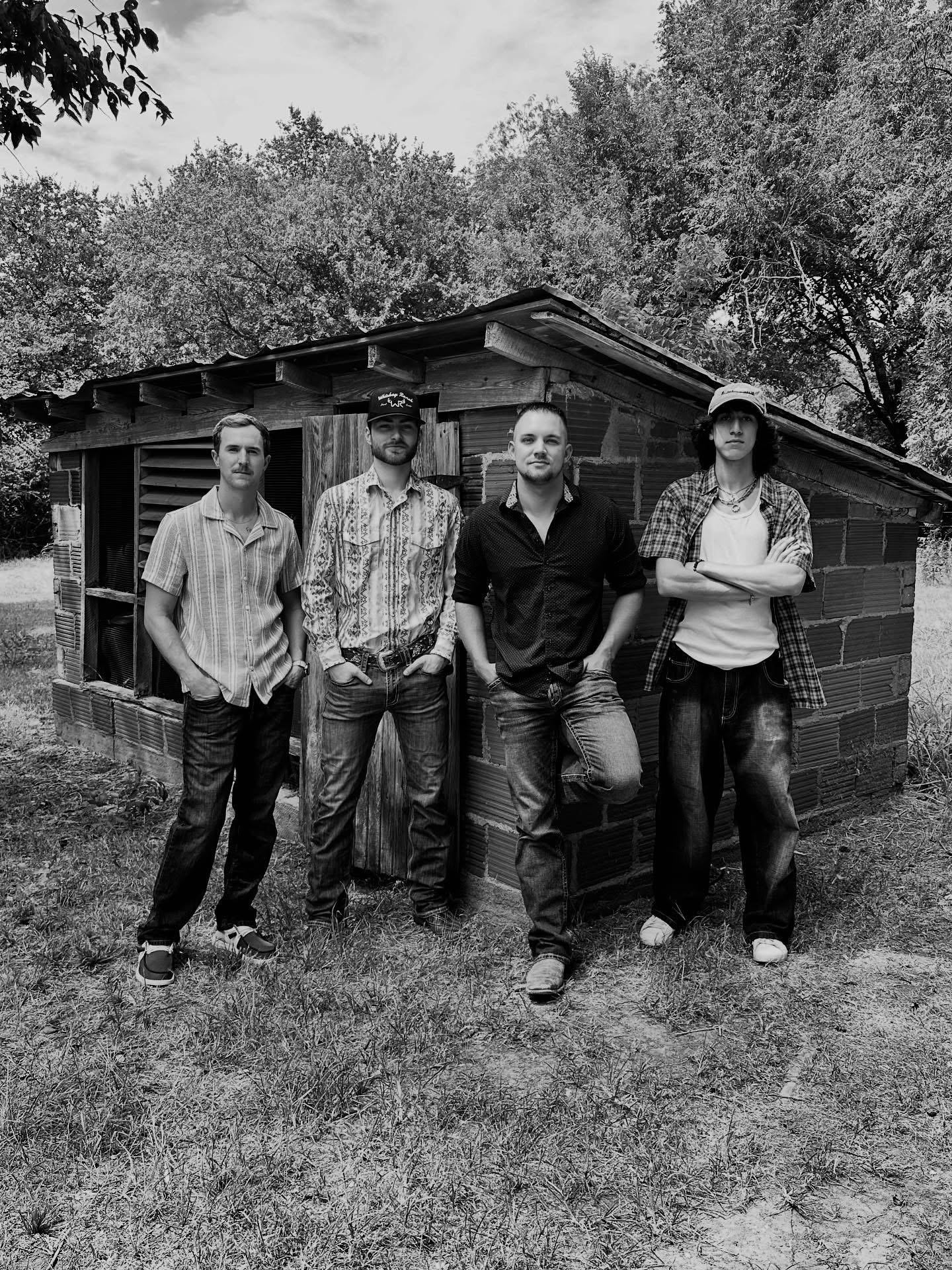 Four young men standing in front of a small, rustic brick and wood building outdoors, with trees in the background, in black and white.