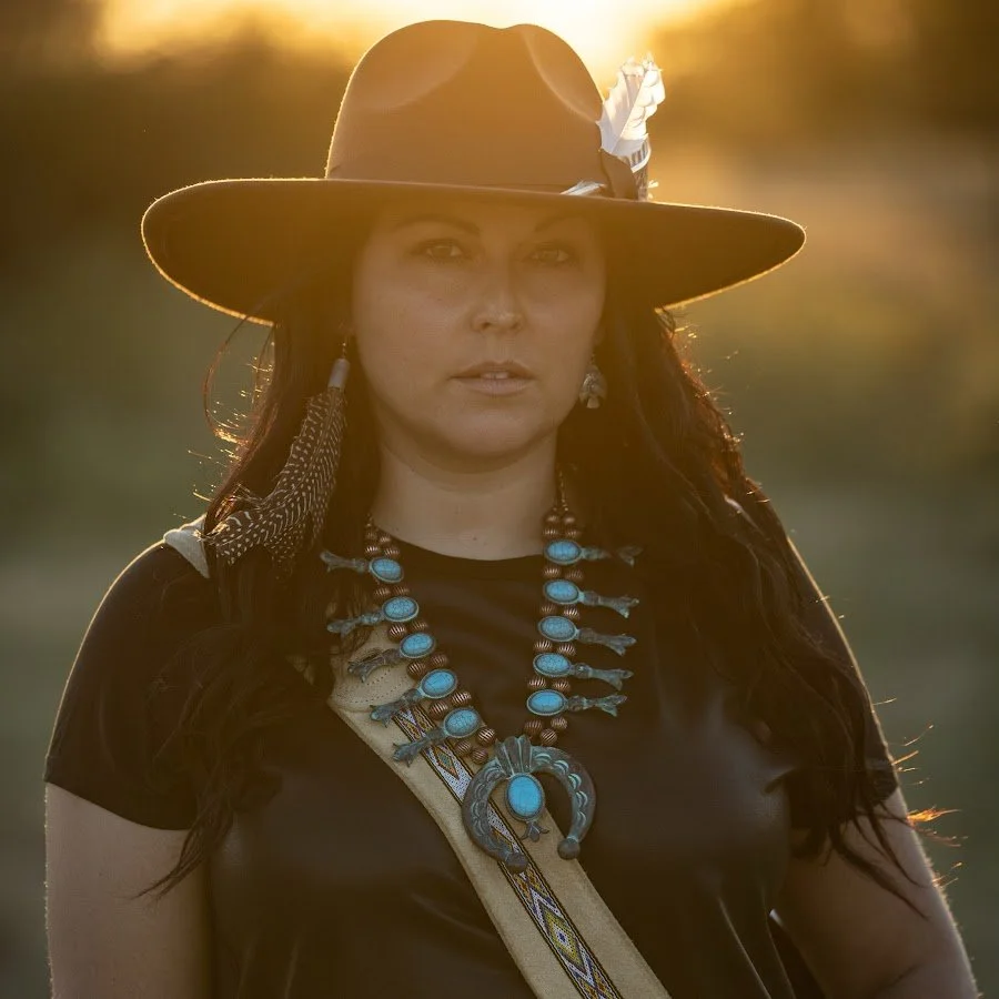 A woman wearing a large floppy hat with a feather, traditional Native American jewelry, and a dark top, standing outdoors at sunset.