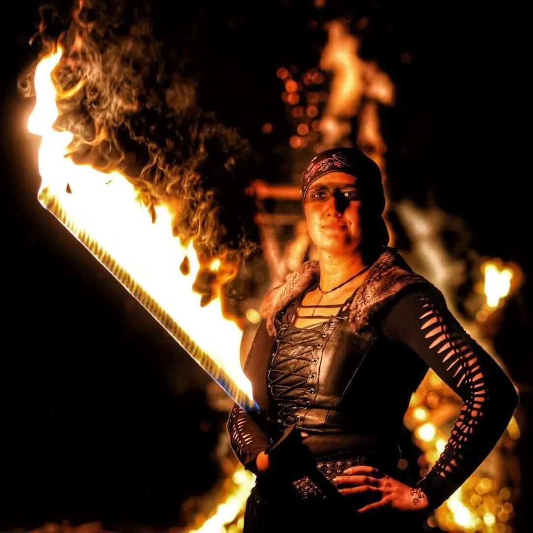 A person wearing a dark, stylized outfit holds a large sword engulfed in flames, standing against a dark background with fire in the distance.