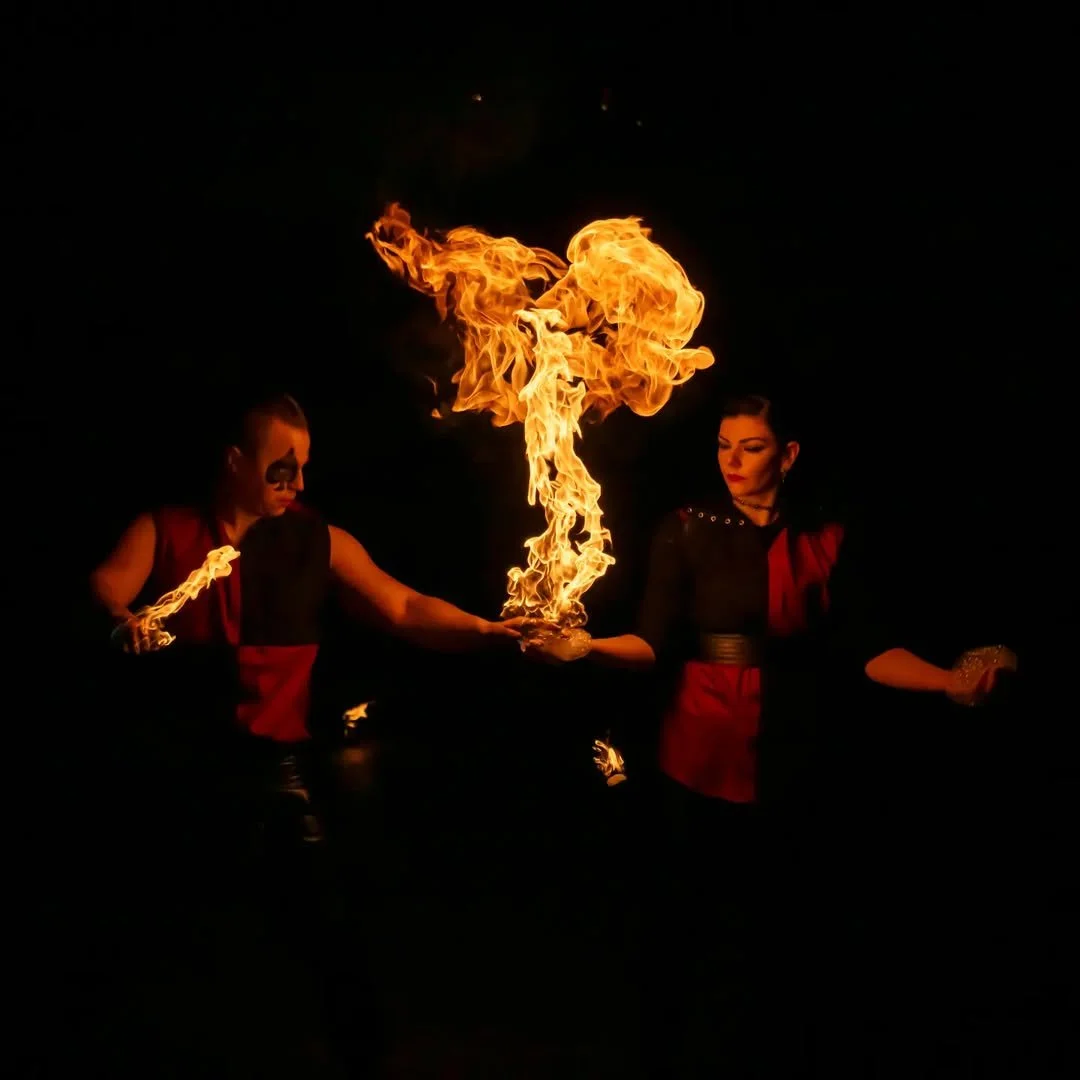 Two performers engaged in a fire show, wearing dark costumes with red accents. One holds a fire torch, while the other appears to pass a flame between their hands, set against a dark background.