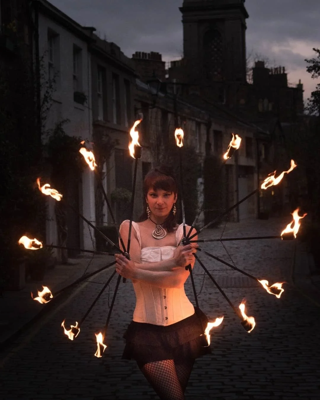 A person performing fire dancing at night on a cobblestone street, holding multiple flaming torches arranged in a circular pattern.