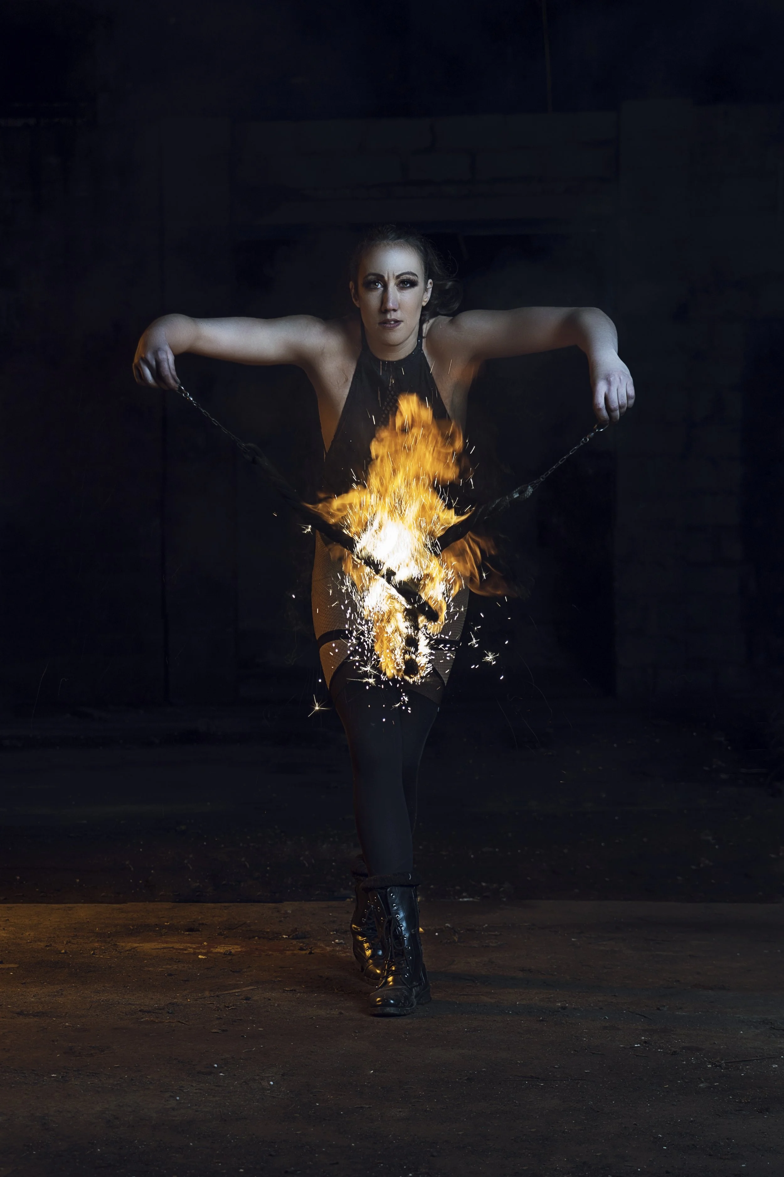 A woman performing a fire dance with flames and sparks, dressed in a black outfit and boots, in a dark setting.