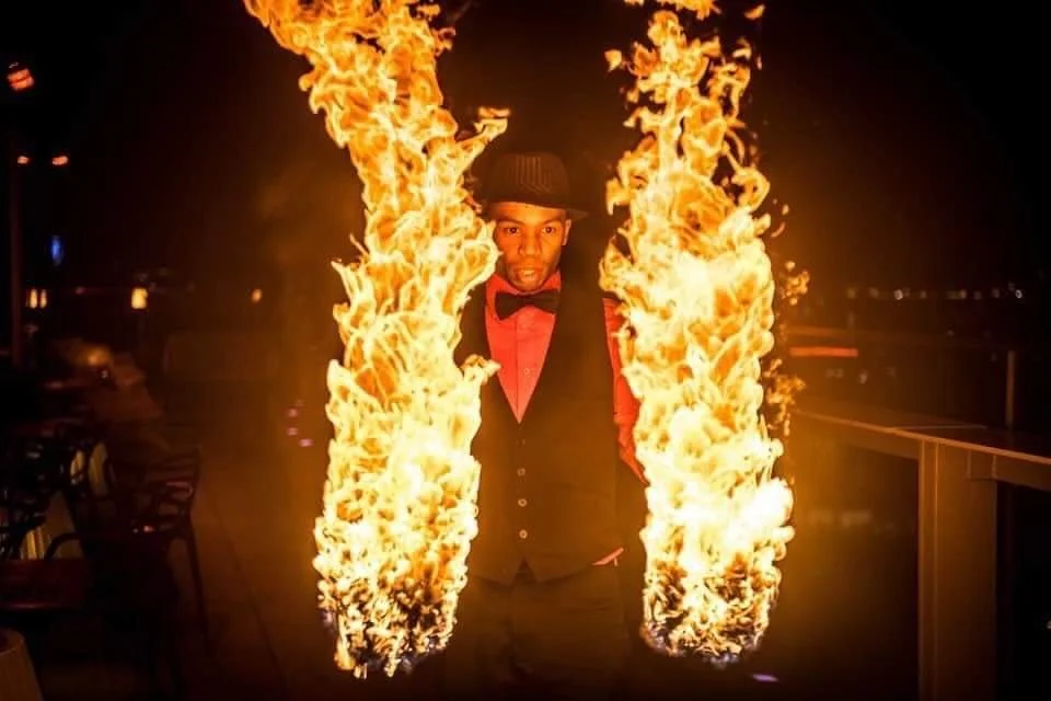 Man performing fire juggling with flaming sticks, wearing a vest, dress shirt, bow tie, and hat at night.
