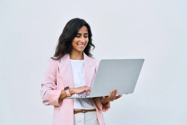 A woman with long dark hair wearing a light pink blazer and white shirt, smiling while holding a silver laptop against a plain white background.