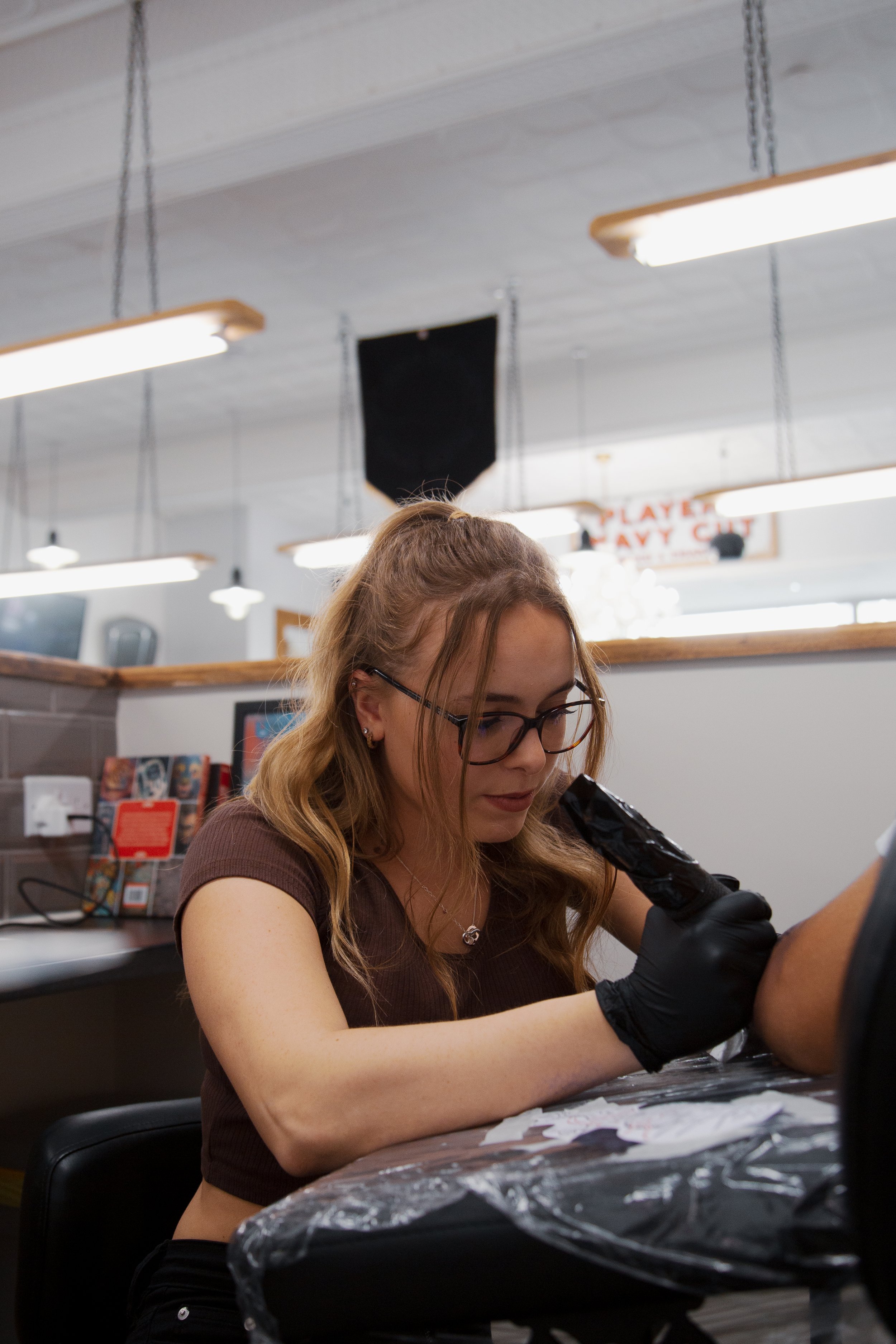 A woman wearing glasses and black gloves tattooing a person's arm in a tattoo studio.