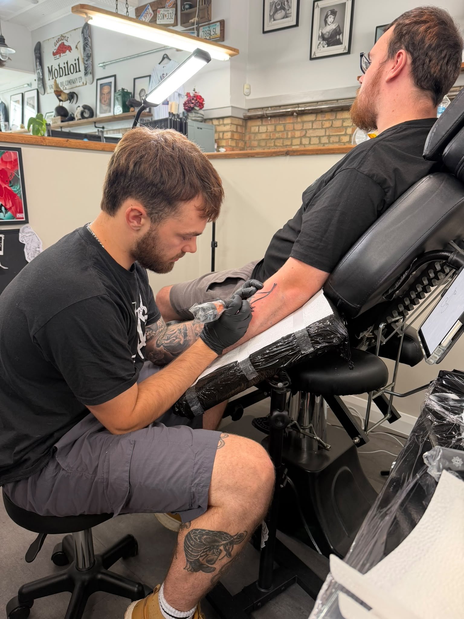A tattoo artist is tattooing a man who is seated in a tattoo studio. The man is wearing a black t-shirt, gray shorts, and yellow work boots. The studio has framed artwork and vintage signs on the walls, and a tattoo machine in use. The artist is focused on his work, tattooing the man's arm.
