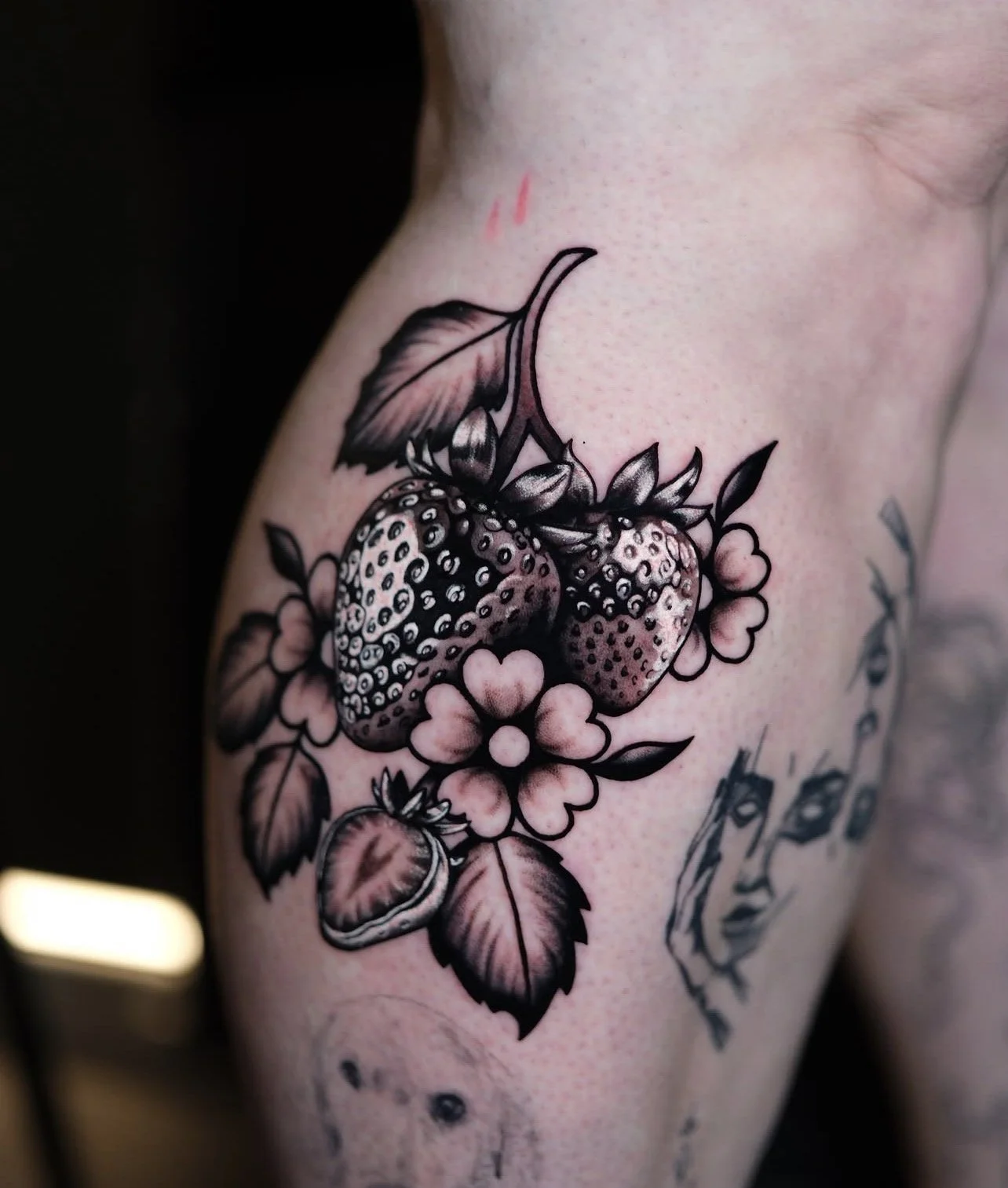 Black and grey chrome hyper-hyper-realisim tattoo of strawberries and flowers on a person's leg.
