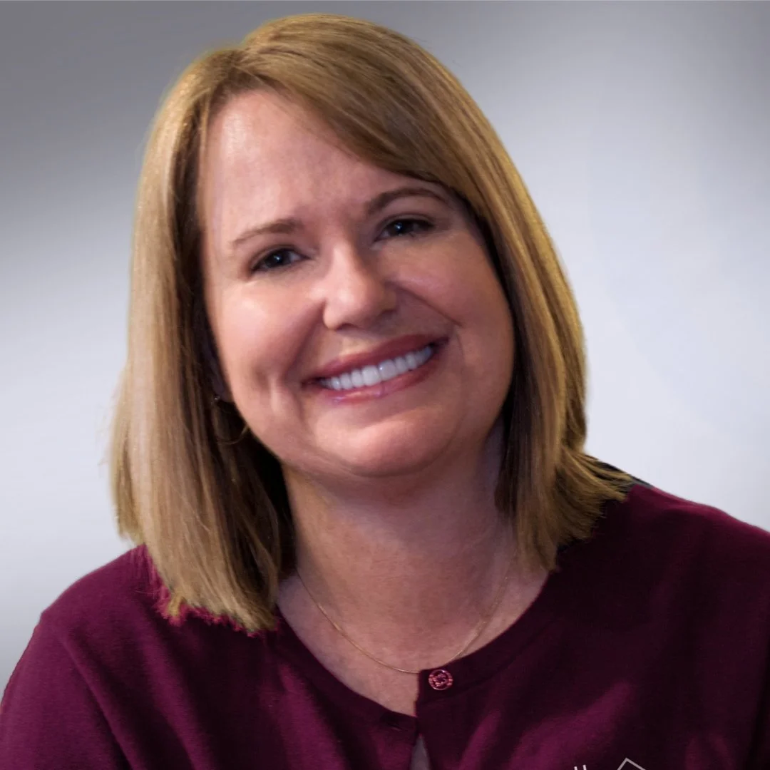 Portrait of a Carleen with shoulder-length light brown hair, smiling, wearing a burgundy top, and a gold necklace.