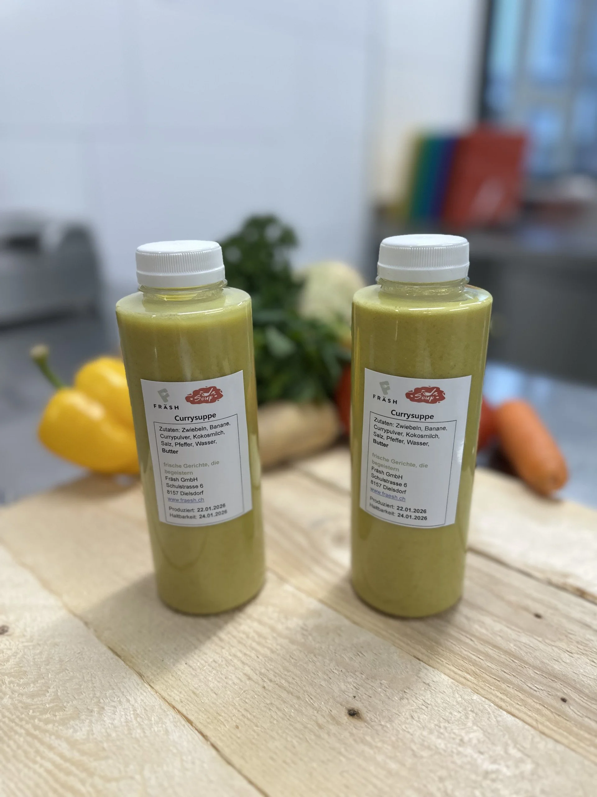 Zwei Flaschen mit grüner Currysoße stehen auf einem Holztisch, im Hintergrund sind Paprika, Knollensellerie und andere Gemüsesorten sichtbar.